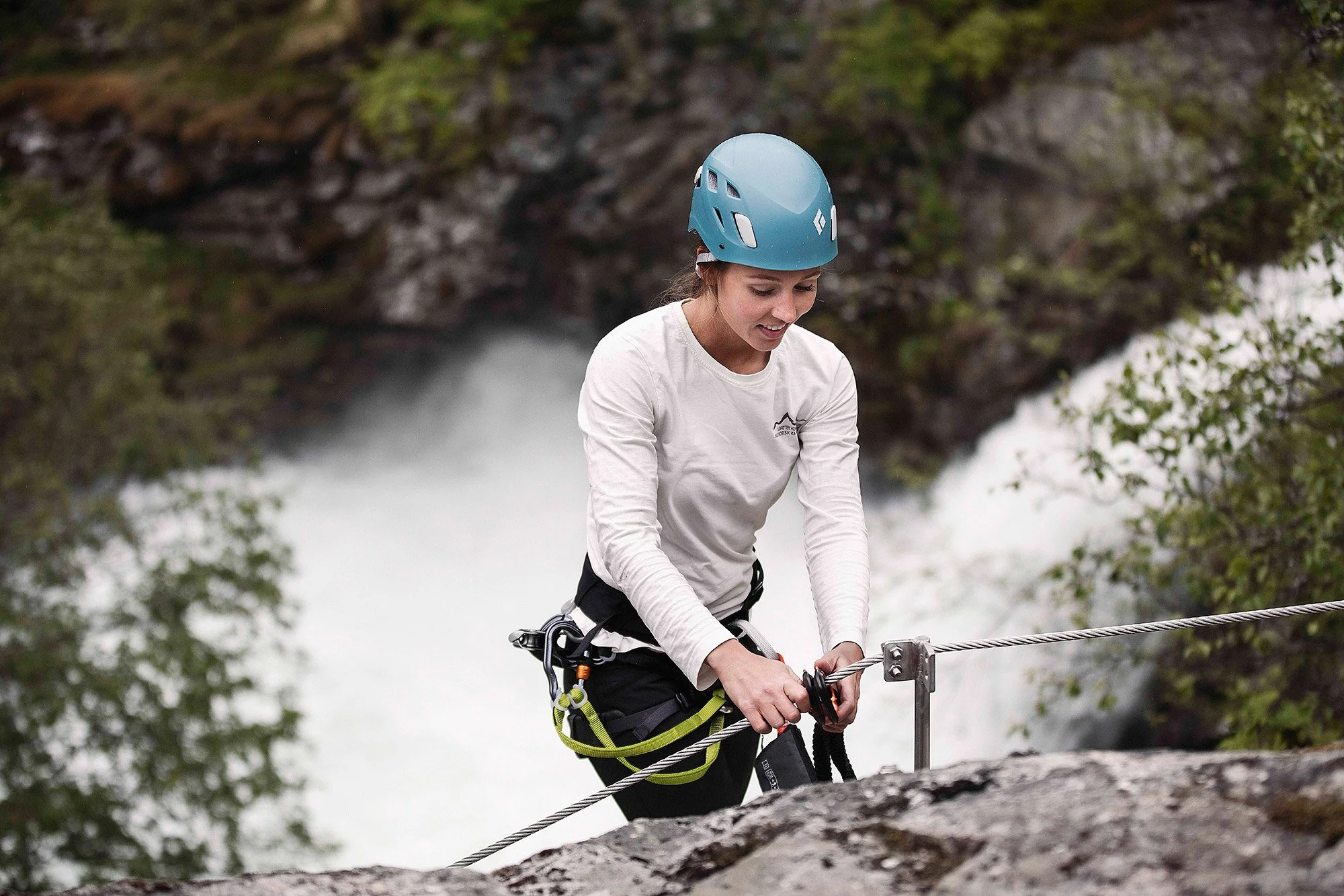 A woman wearing a helmet and climbing gear, holding a safety rope, standing near a waterfall, surrounded by trees.