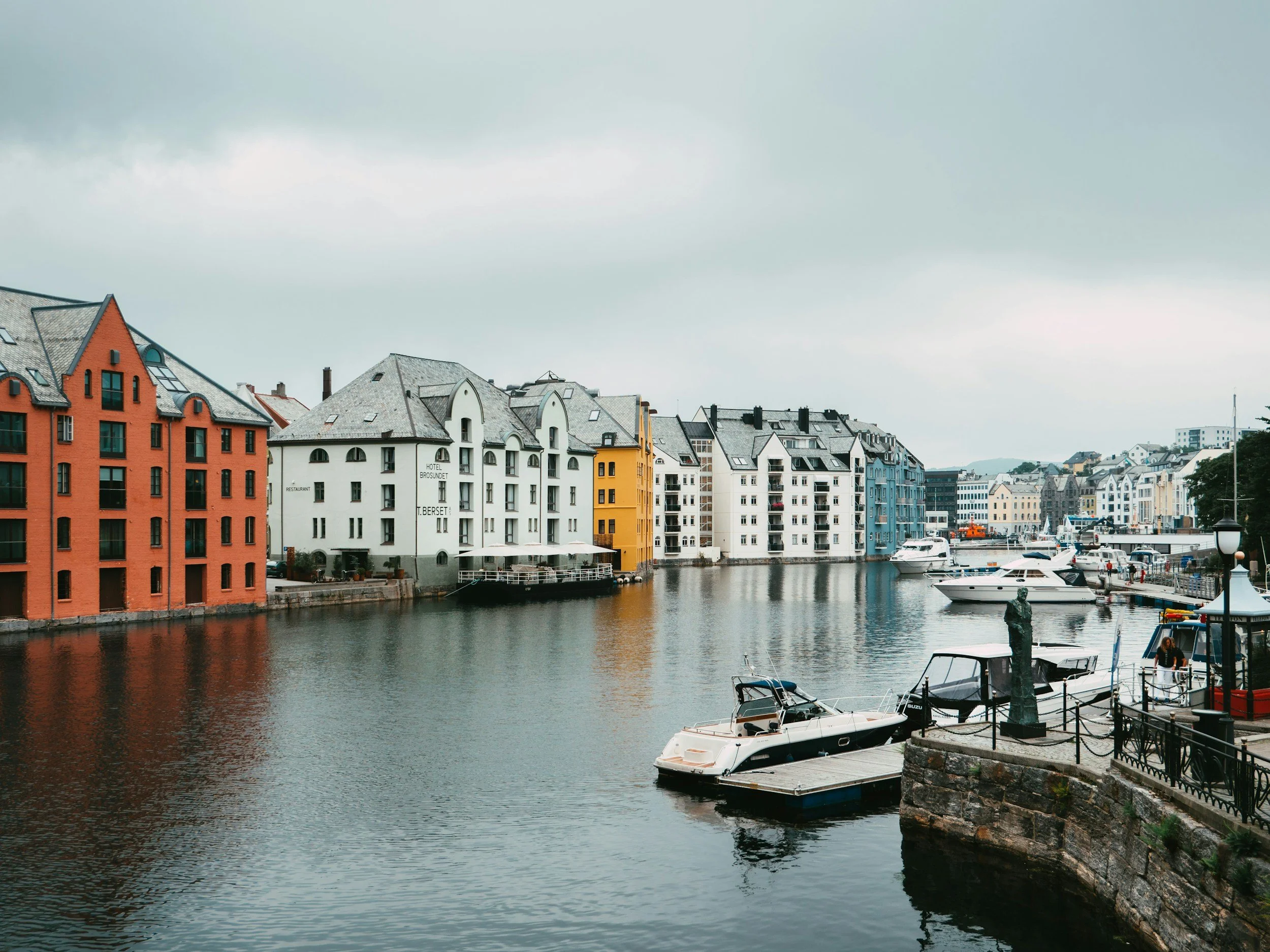 Explore Norway. ålesund. Colorful buildings along a harbor with boats docked on calm water and cloudy sky overhead.