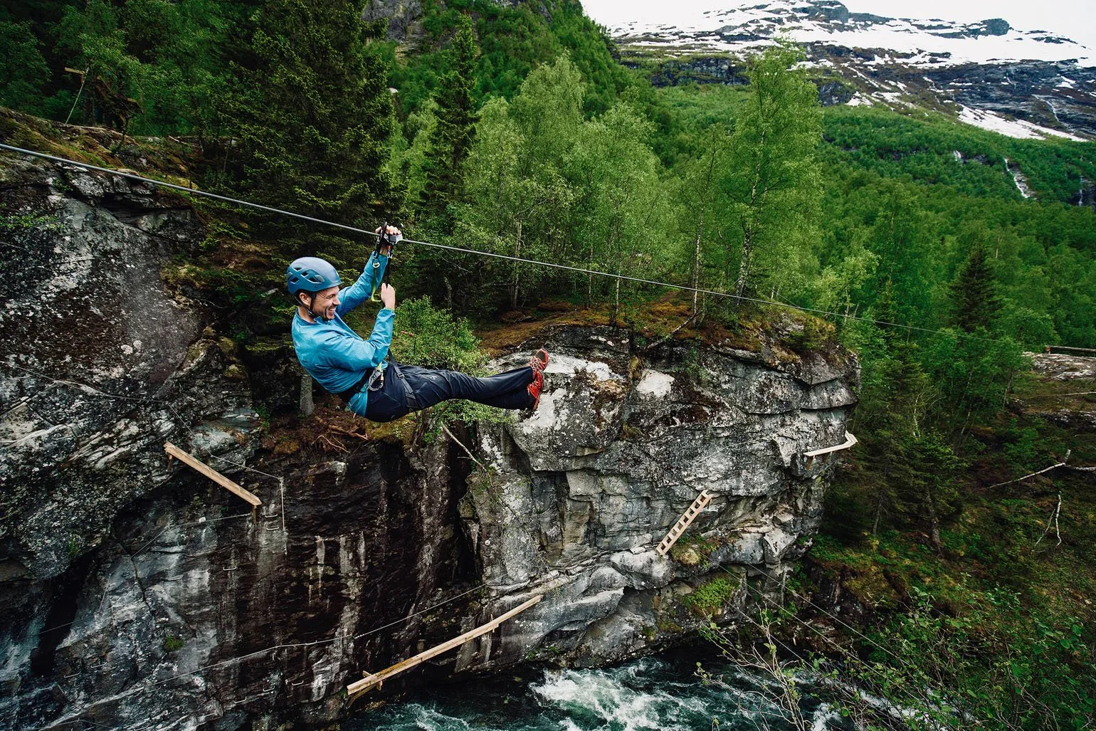 Person in blue jacket and helmet ziplining over a river in a forested mountainous area.