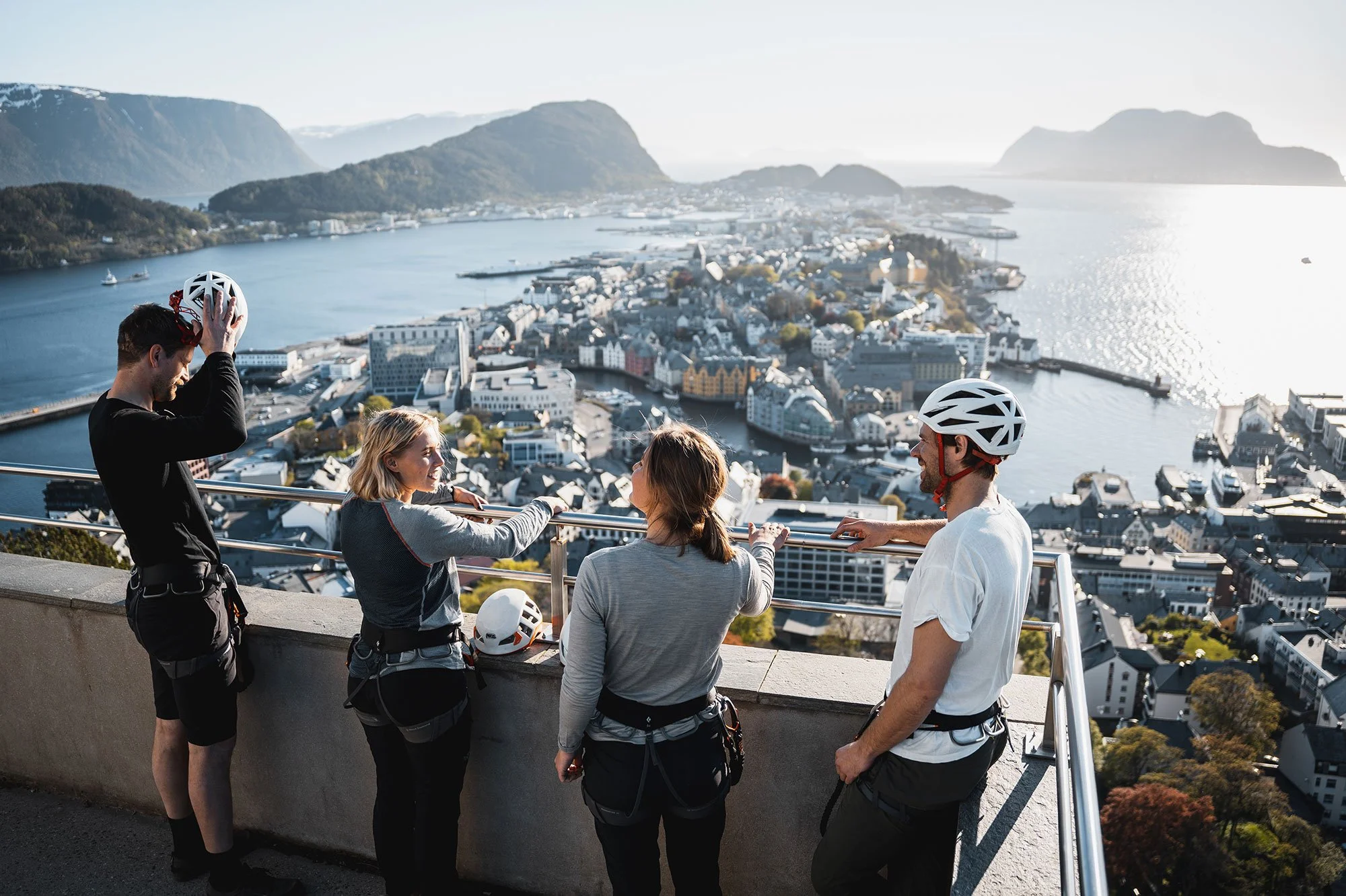 Explore Norway. Four people in helmets and outdoor clothing standing on a viewing platform overlooking a coastal city with water and mountains in the background, engaging in a conversation.