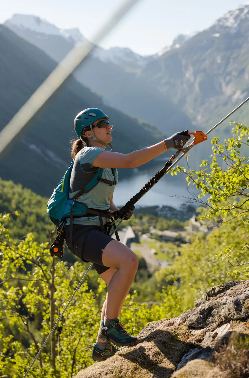 Explore Norway.A woman rock climbing outdoors in a scenic mountainous area with greenery and snow-capped peaks in the background. She wears a helmet, sunglasses, a harness, and athletic clothing, and is using a rope and carabiner to assist her climb.