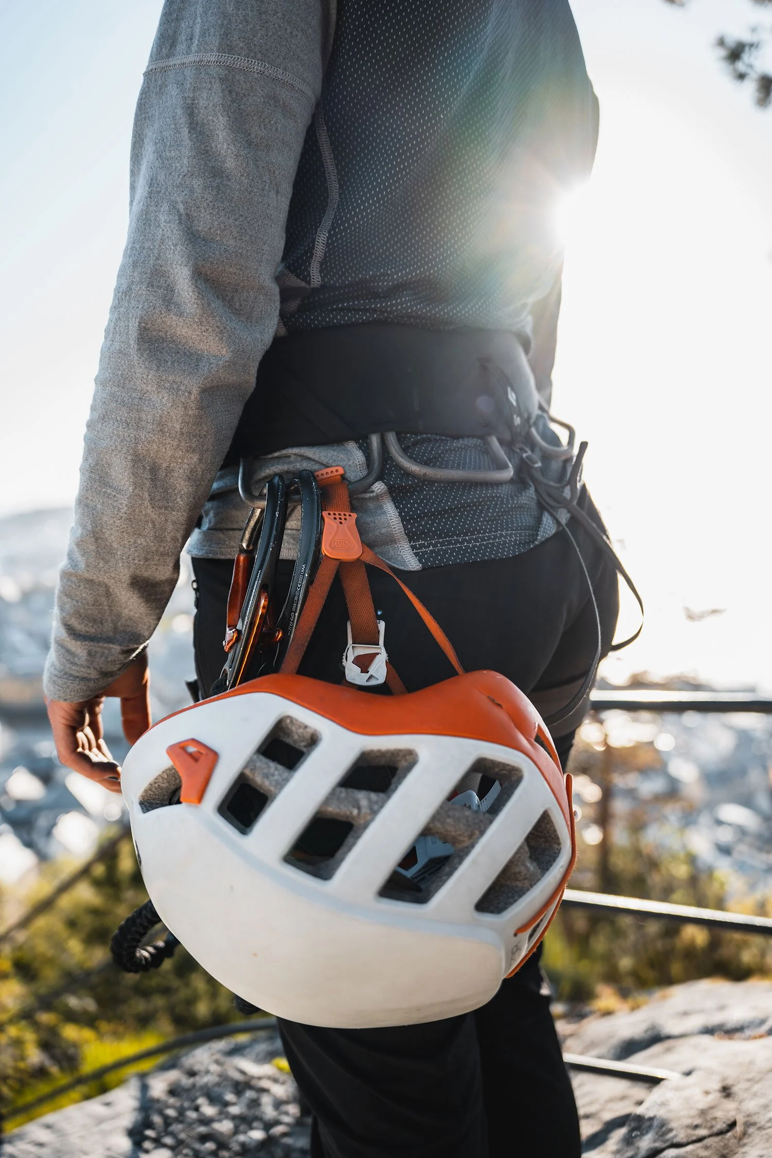 Explore Norway. Close-up of a person wearing outdoor gear, holding a climbing helmet with orange and white colors, with carabiners attached to their waist, outdoors with sunlight and nature in the background.