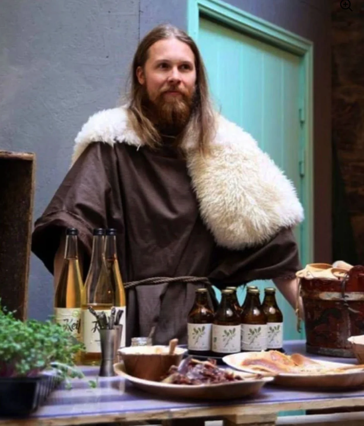 A man dressed in medieval or fantasy-style clothing with a brown robe and a white fur stole, standing behind a table with food and drinks, indoors with wooden and colorful walls.