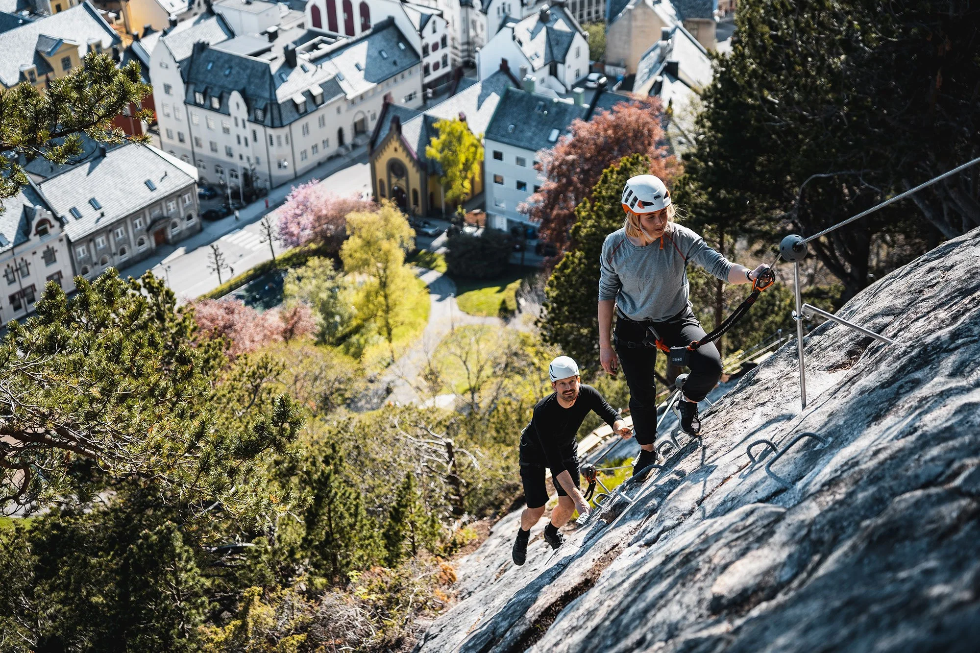 Explore Norway. Two people rock climbing on a steep outdoor climbing wall, wearing helmets and climbing gear, with a cityscape and trees in the background.