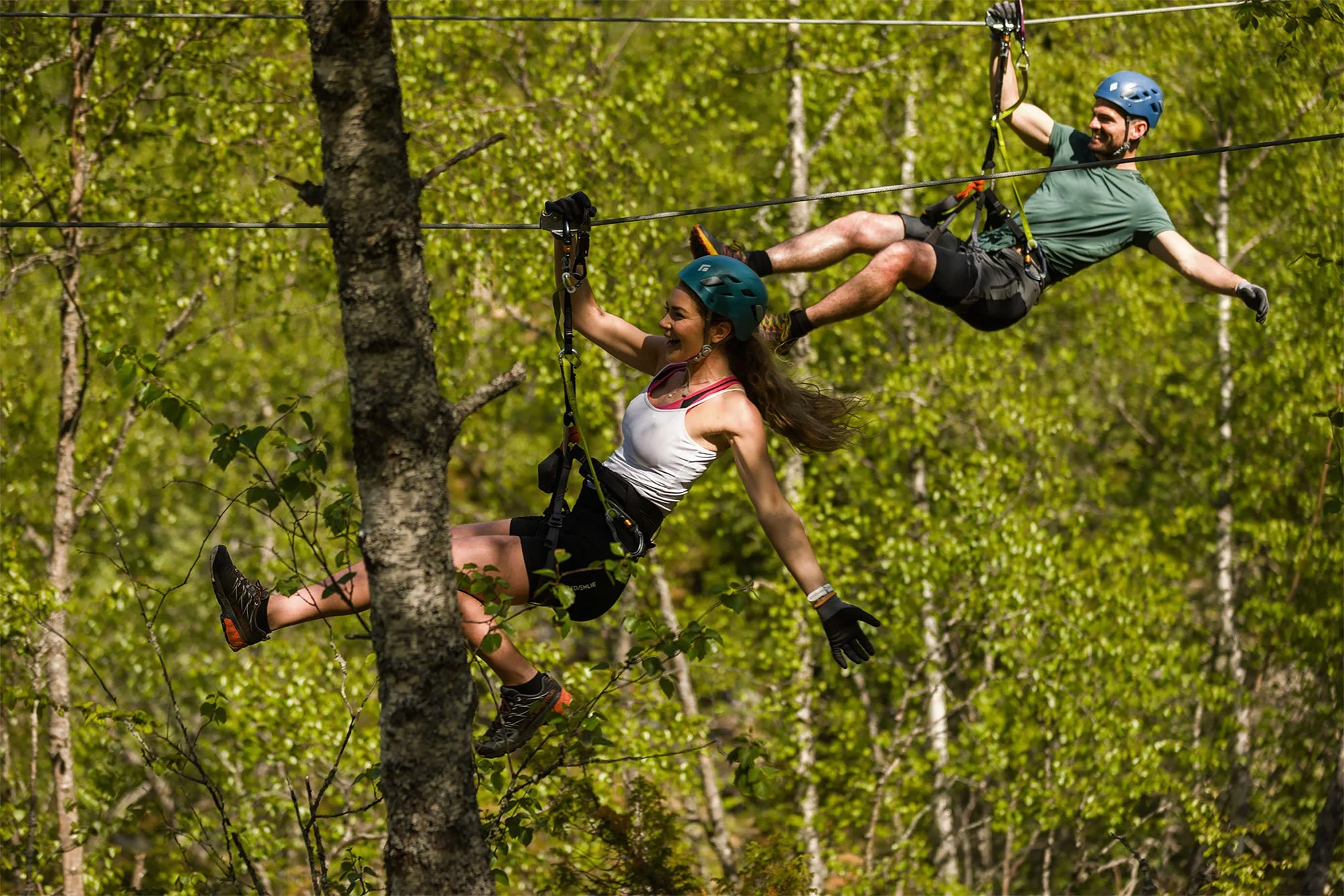 Two people zip lining through a forest with green trees, one man and one woman both wearing helmets and harnesses.