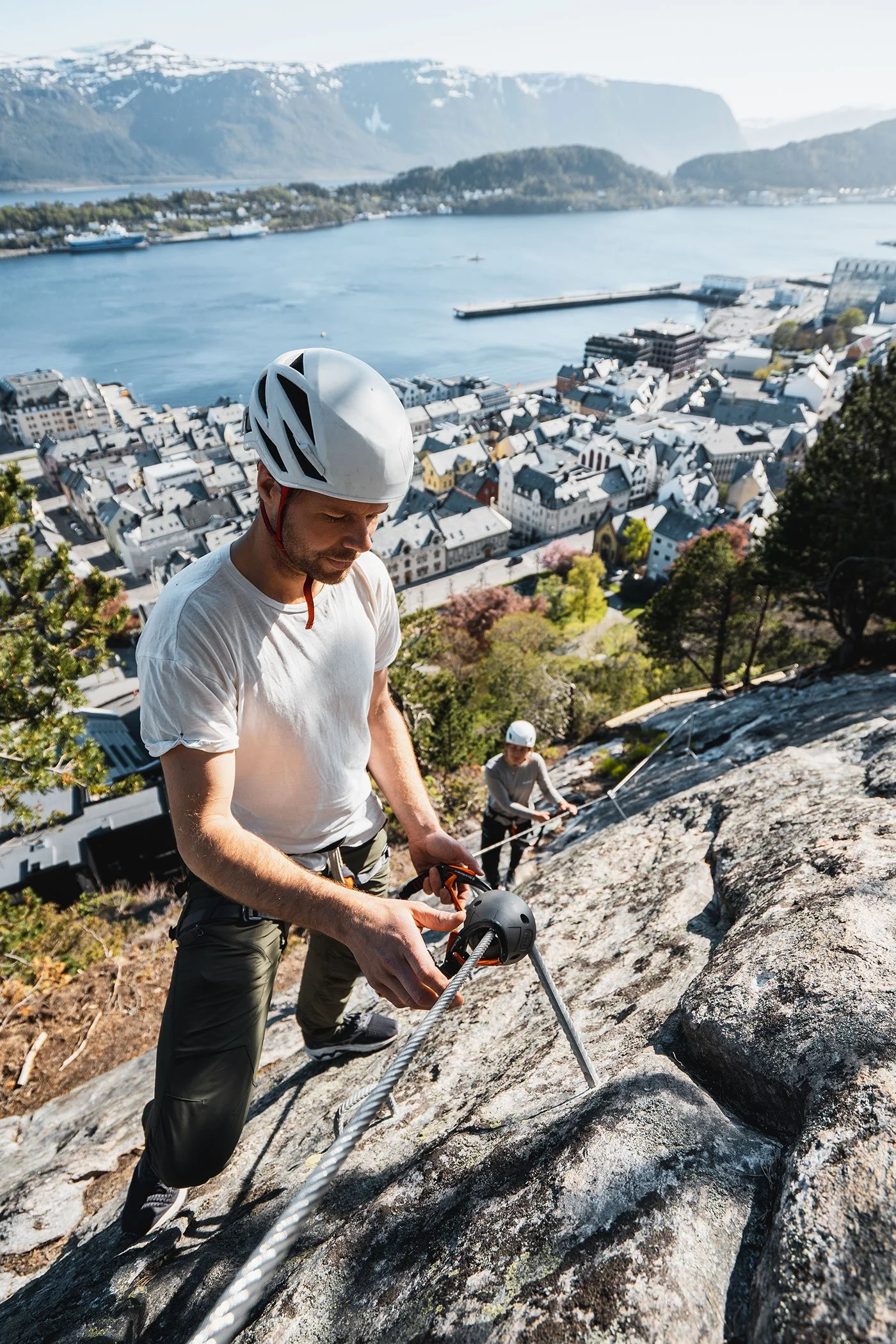 Man wearing a white helmet and white t-shirt climbing a steep rock face using a safety rope, with a scenic town, water, mountains, and another climber in the background.