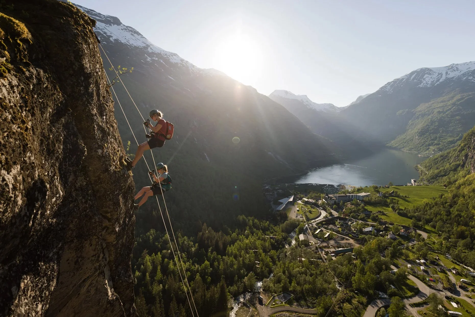 Two people climbing a steep cliff face in a mountainous landscape with a lake and town below, during daytime.