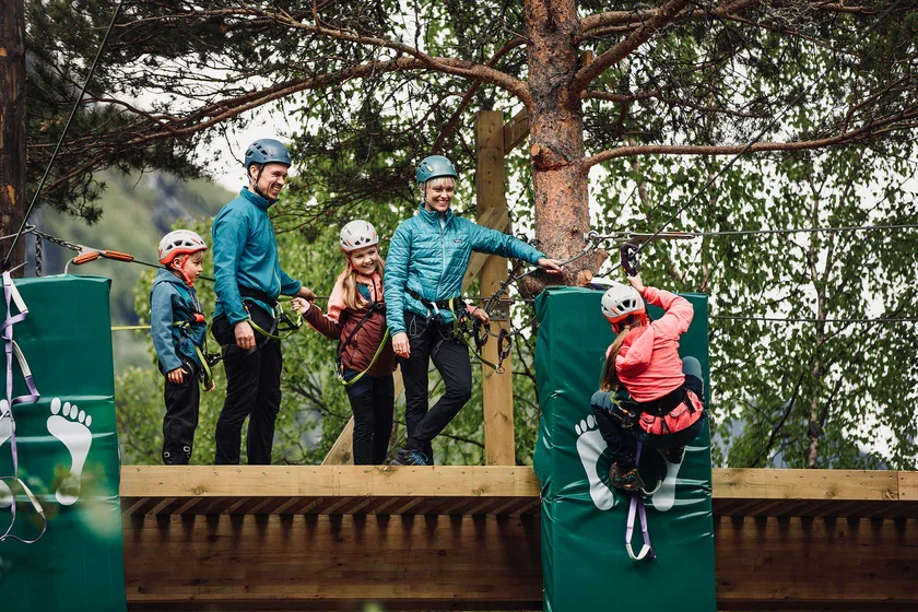 A group of five people, including children and adults, participating in a tree-top adventure course, all wearing helmets and harnesses, with one adult assisting a child climbing a wall.