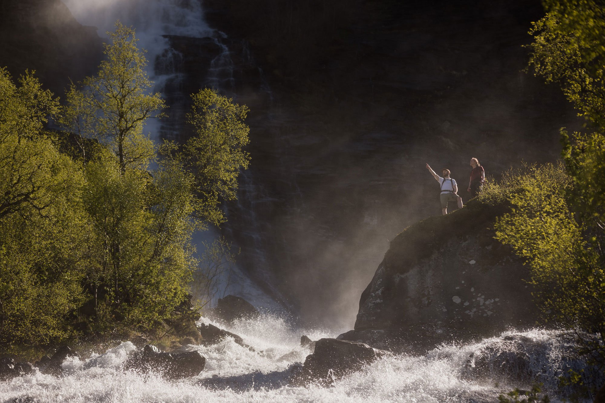 Two hikers with backpacks standing on a rocky ledge, overlooking a waterfall in a lush green forest.