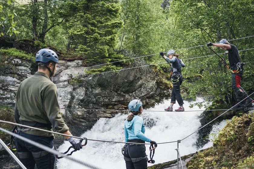 Four people wearing helmets and harnesses cross a hanging suspension bridge over a rushing waterfall in a forest.