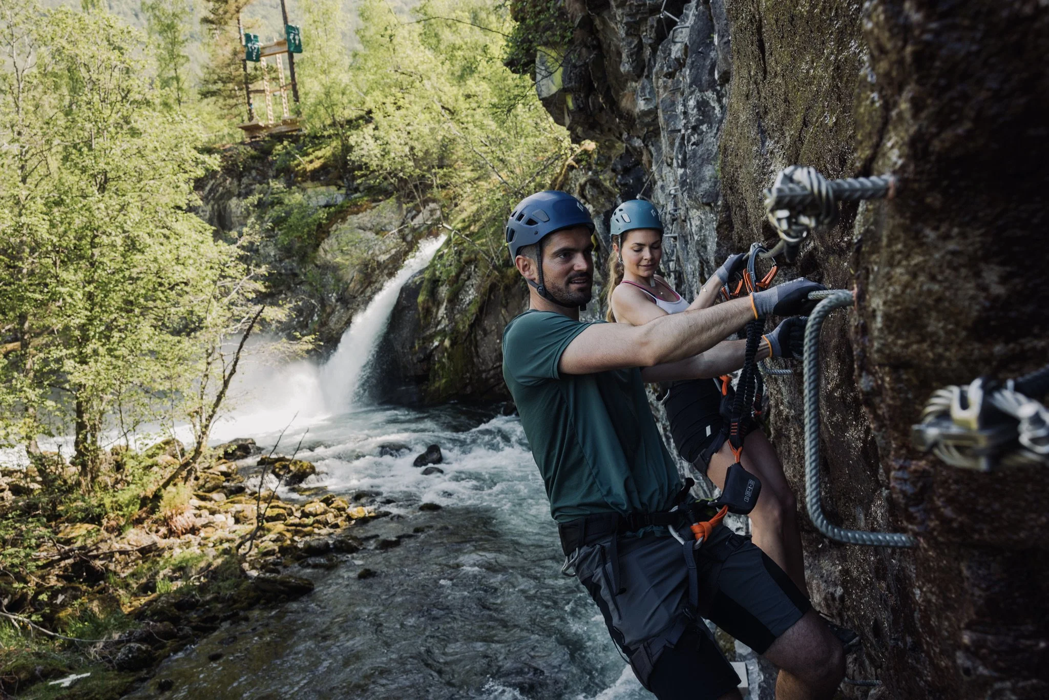 Two people rock climbing on a cliff beside a flowing river in a forested area with a waterfall in the background.