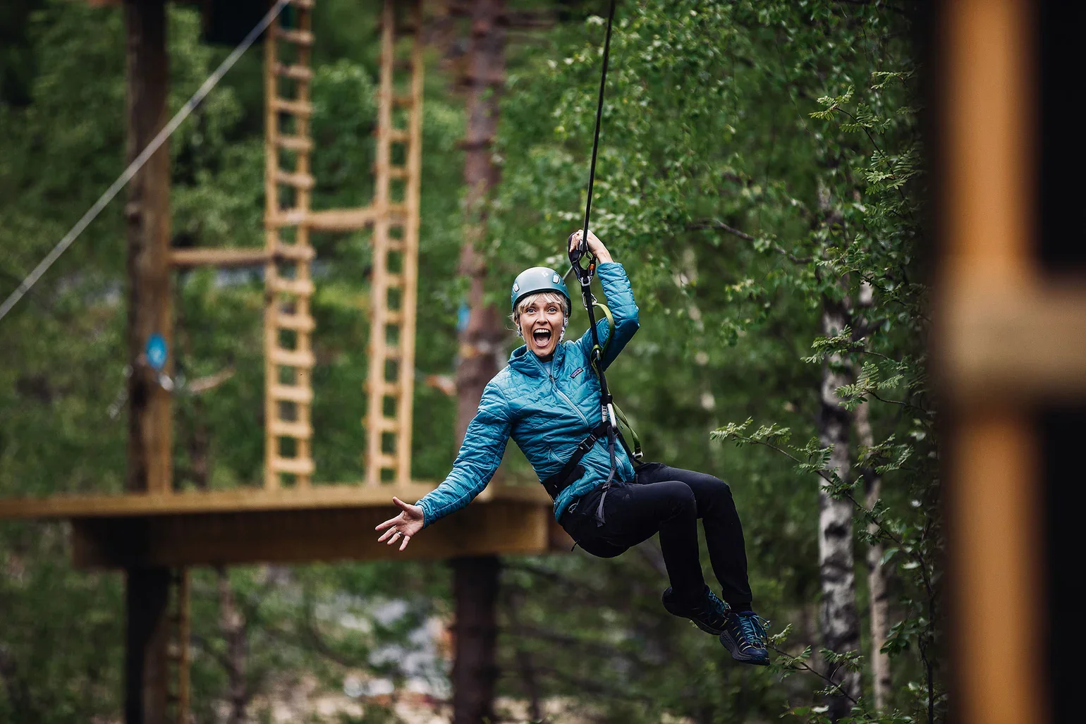 Woman wearing a helmet and blue jacket riding a zip line in a wooded area, smiling.