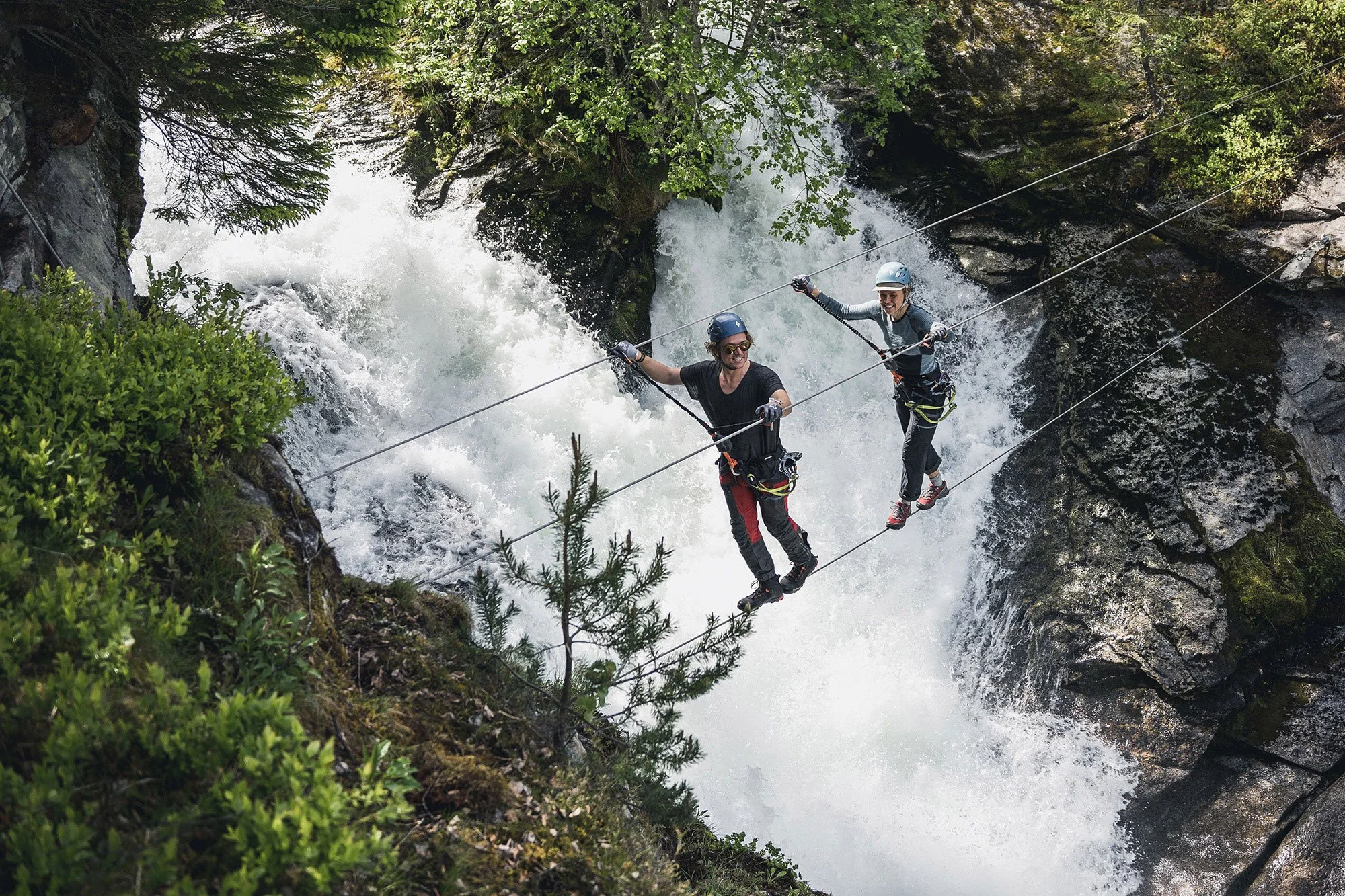 Two people are walking on a high wires suspension bridge over a rushing mountain river with rocks and lush greenery.