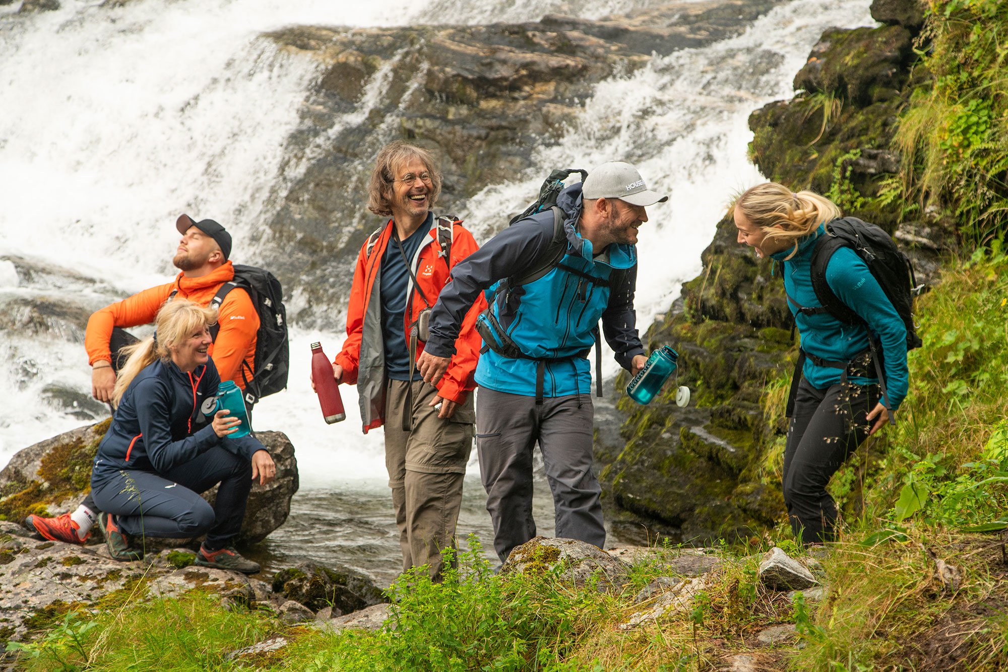 Group of five hikers, three men and two women, standing on rocks near a waterfall, smiling and laughing, wearing outdoor gear and backpacks.
