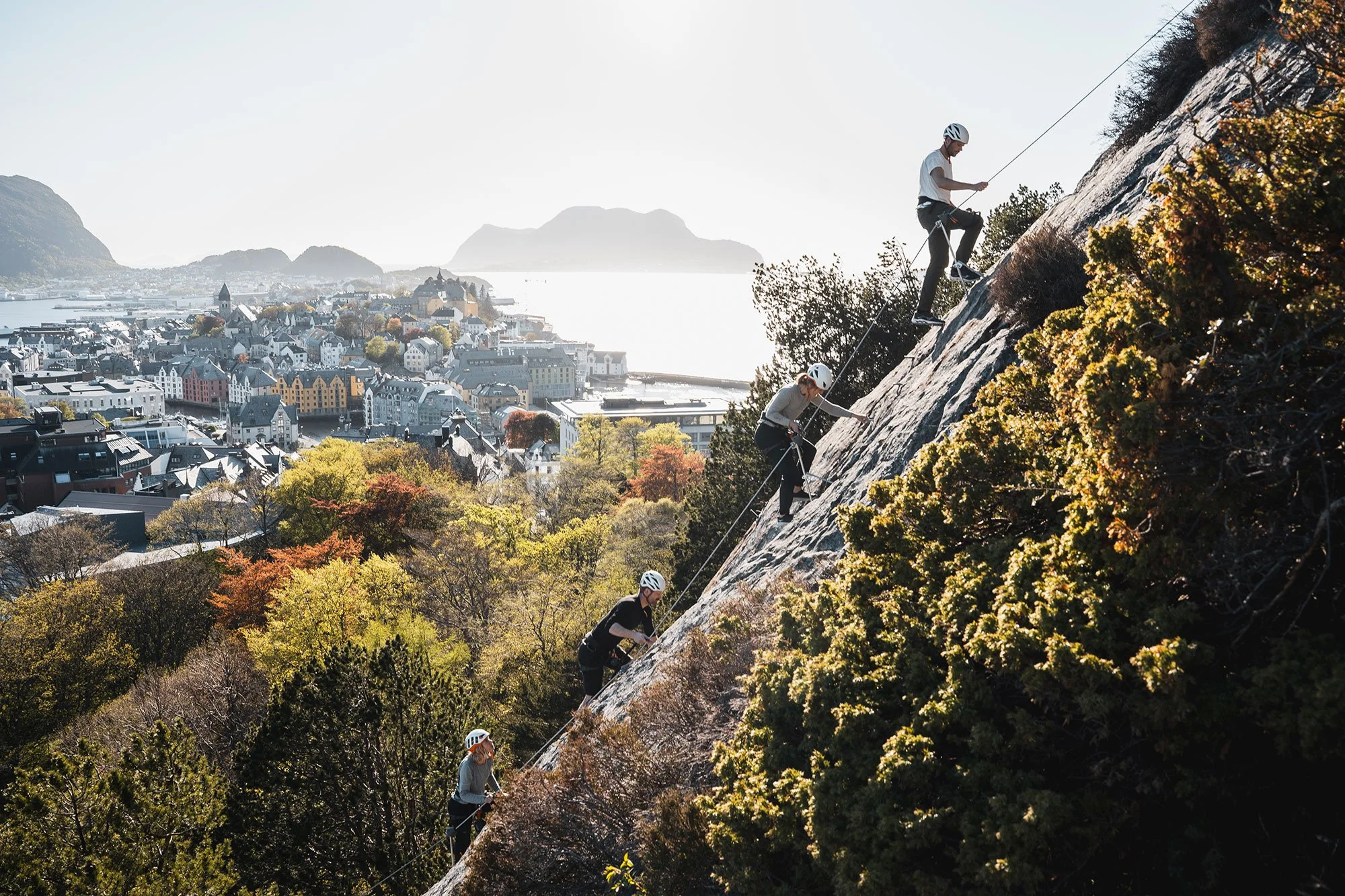 Explore Norway. Ålesund. Group of five rock climbers ascending a steep rock face on a hillside with a city and water in the background, all wearing helmets.