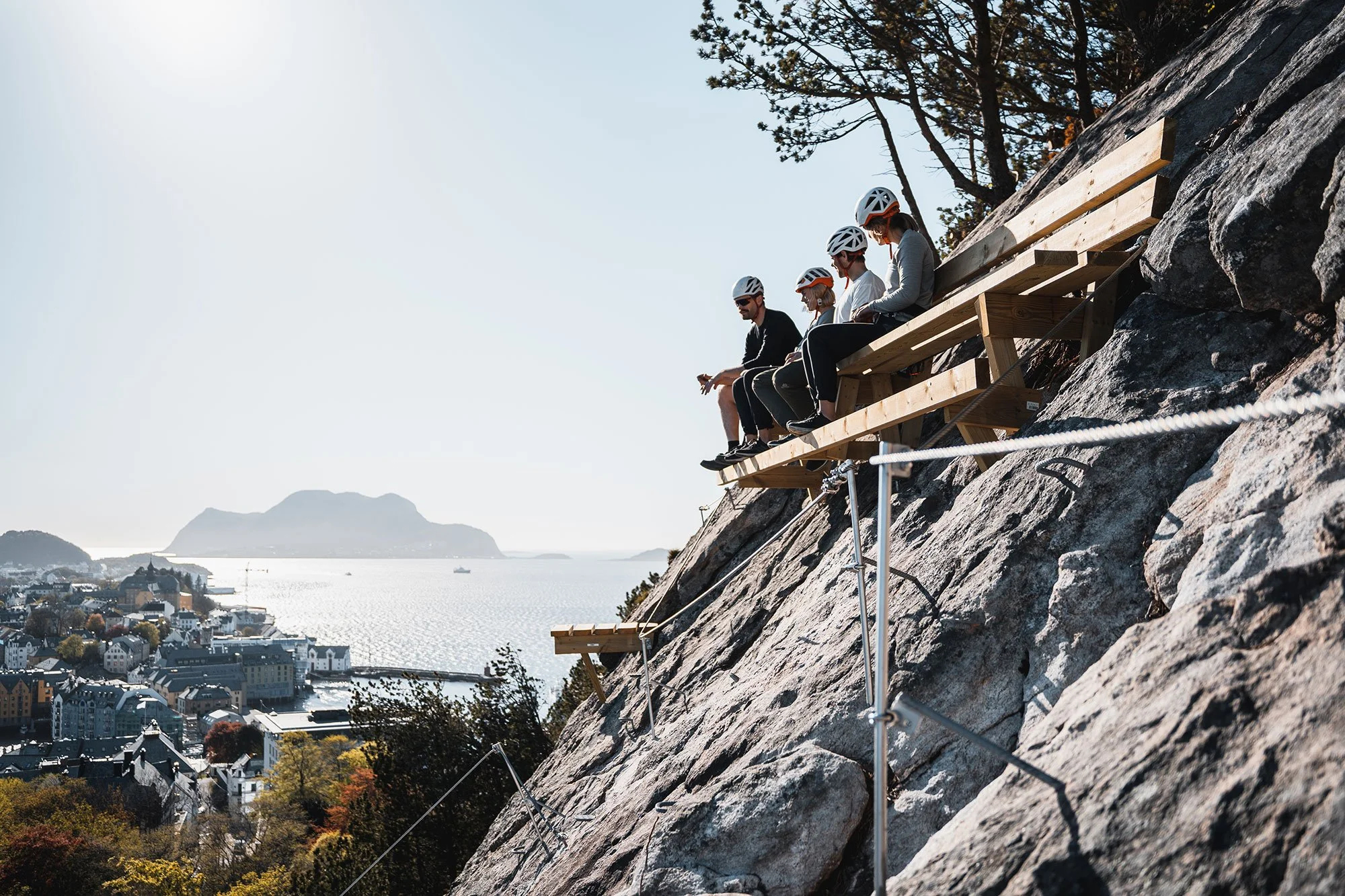 Explore Norway. Ålesund. Five people wearing helmets sit on a wooden platform on a rocky cliff, overlooking a city and body of water.