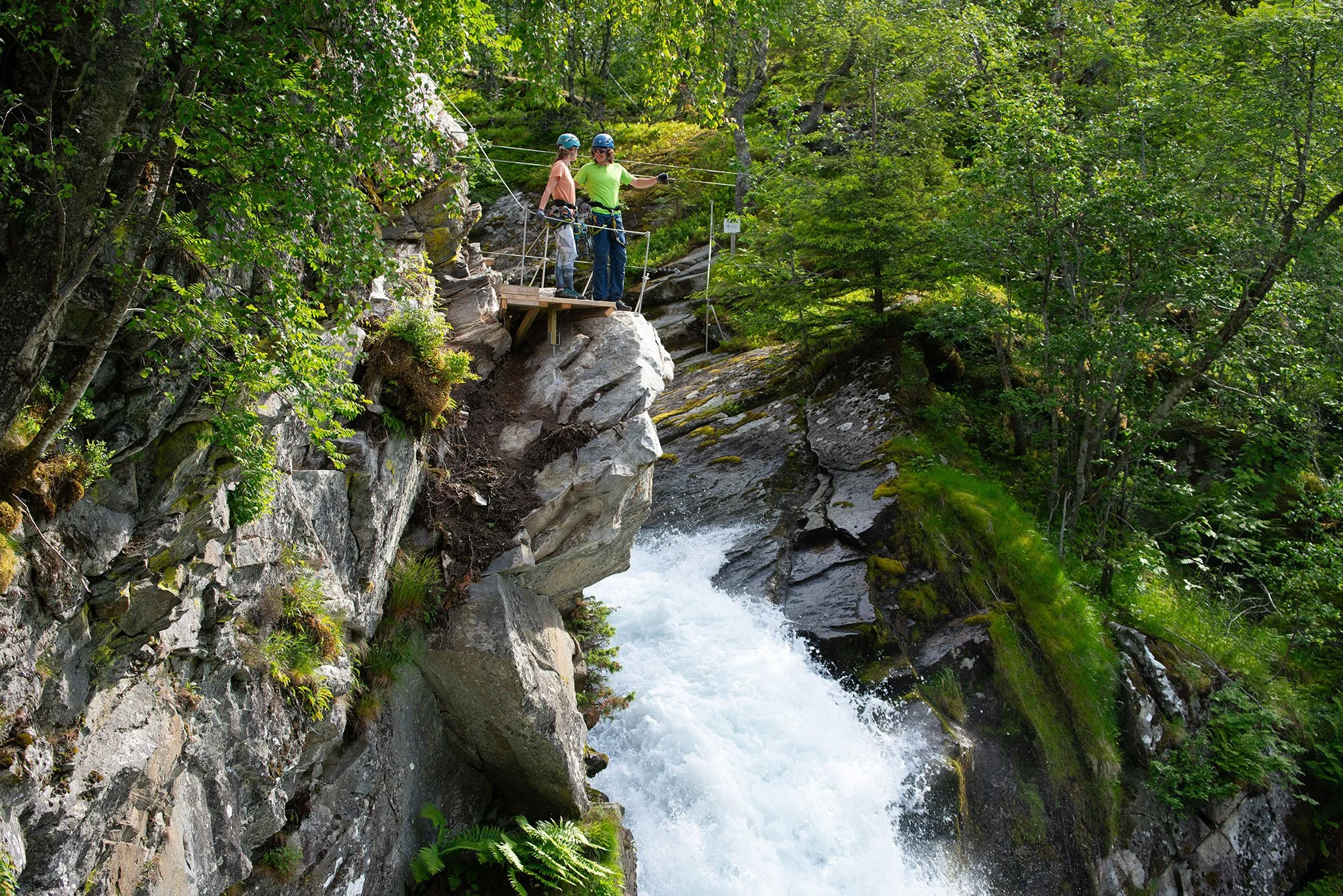 Two people in helmets and harnesses standing on a small wooden platform beside a waterfall, preparing for a canyoning adventure in a lush forest.