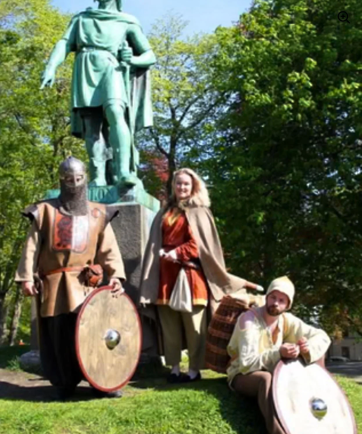 Three individuals dressed in medieval costumes standing and sitting in front of a large green statue of a historical figure outdoors surrounded by trees.