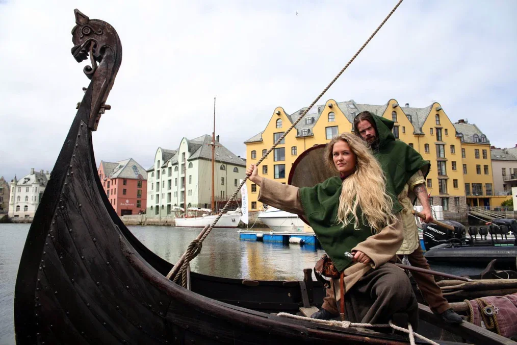 Explore Norway. Two people dressed in Viking-style clothing on a Viking boat with a dragon-shaped prow, on a river with colorful buildings in the background.