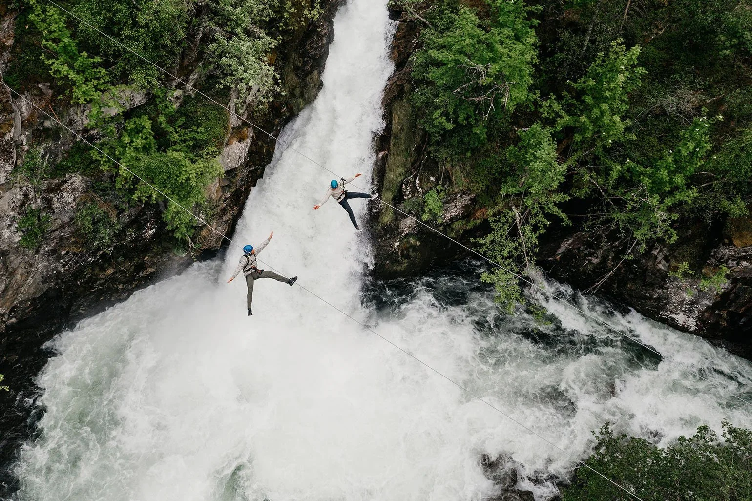 Two people are ziplining across a fast-moving river with a waterfall in the background. They are wearing helmets and safety harnesses, and are surrounded by green trees and rocky cliffs.