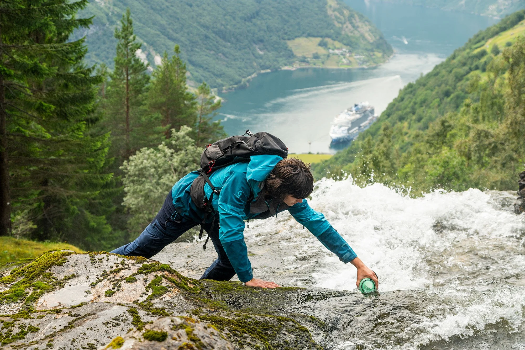Person in a blue jacket and backpack climbing a wet rocky ledge above a river with a cruise ship in the distance, surrounded by green forested mountains.
