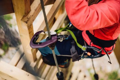 Person in a red jacket working on a wooden structure, using climbing or safety equipment.