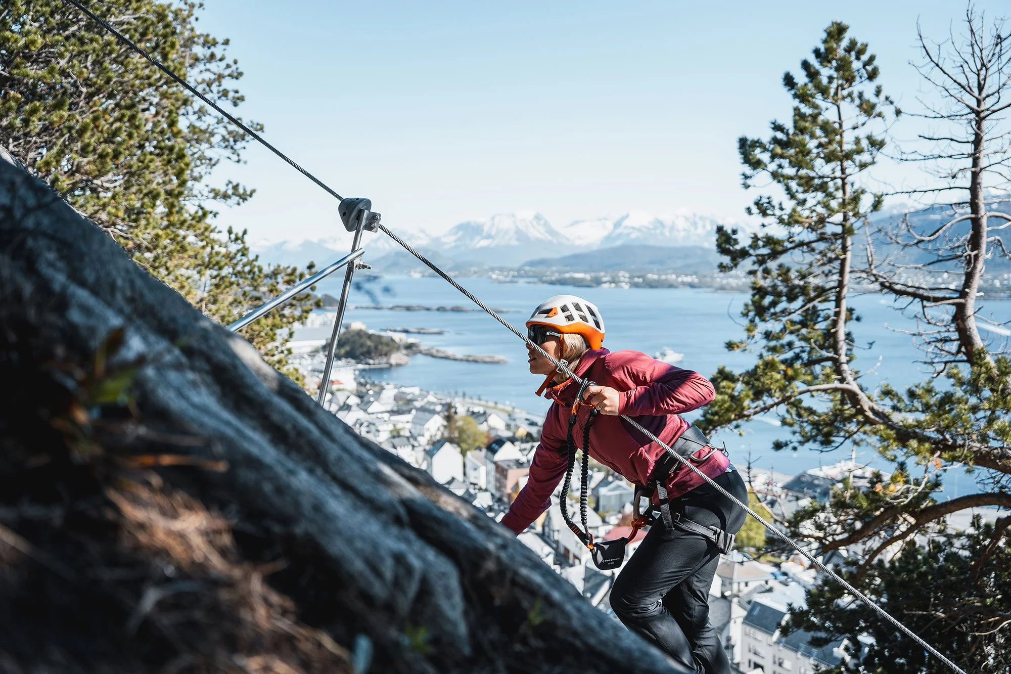 A woman rock climbing on a steep outdoor cliff, wearing a helmet, harness, and a red jacket. In the background, there's a body of water, mountains, and a town with white buildings by the water. Pine trees frame the scene.