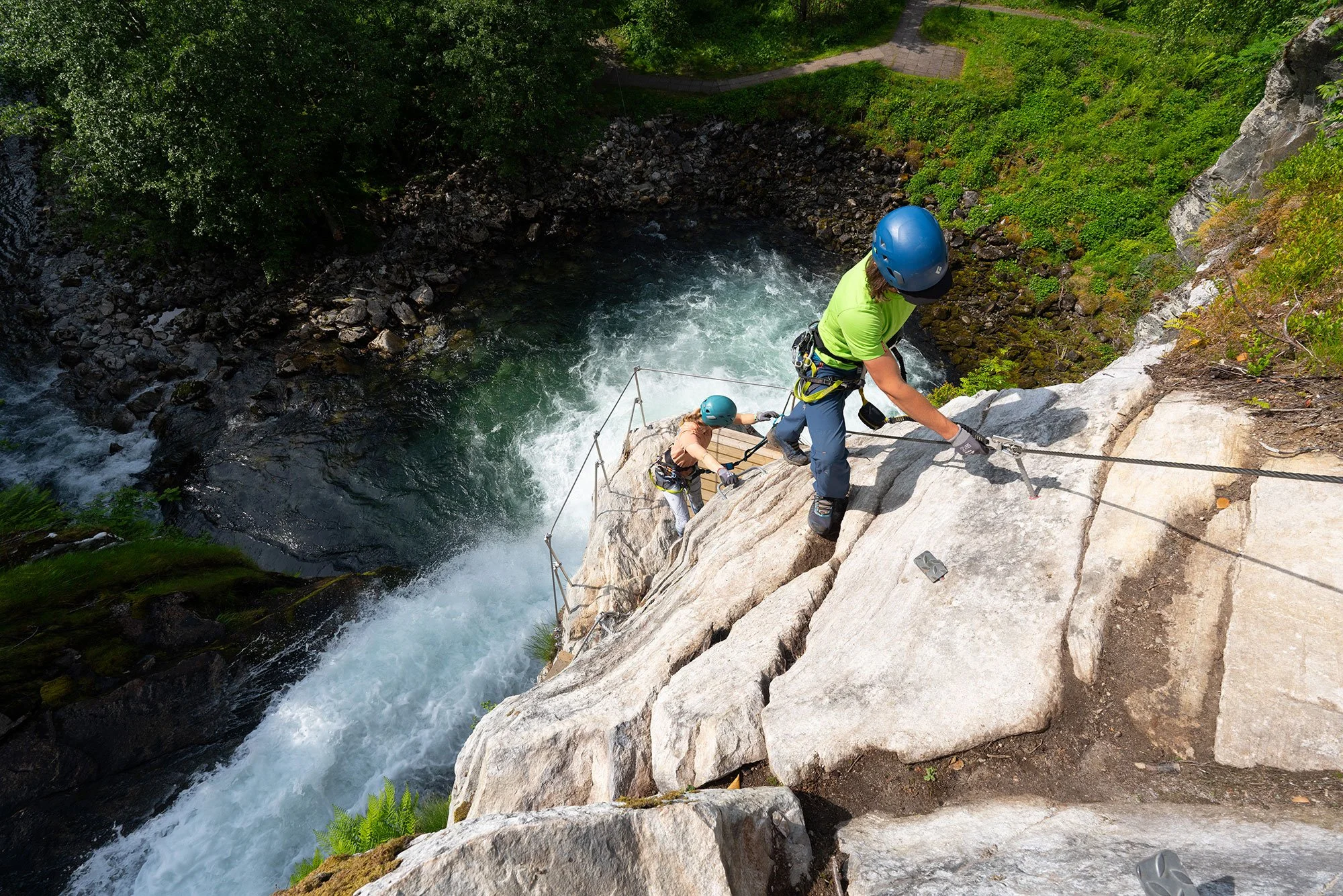 Two rock climbers wearing helmets and harnesses ascending a steep cliff over a river, with grassy and wooded surroundings.