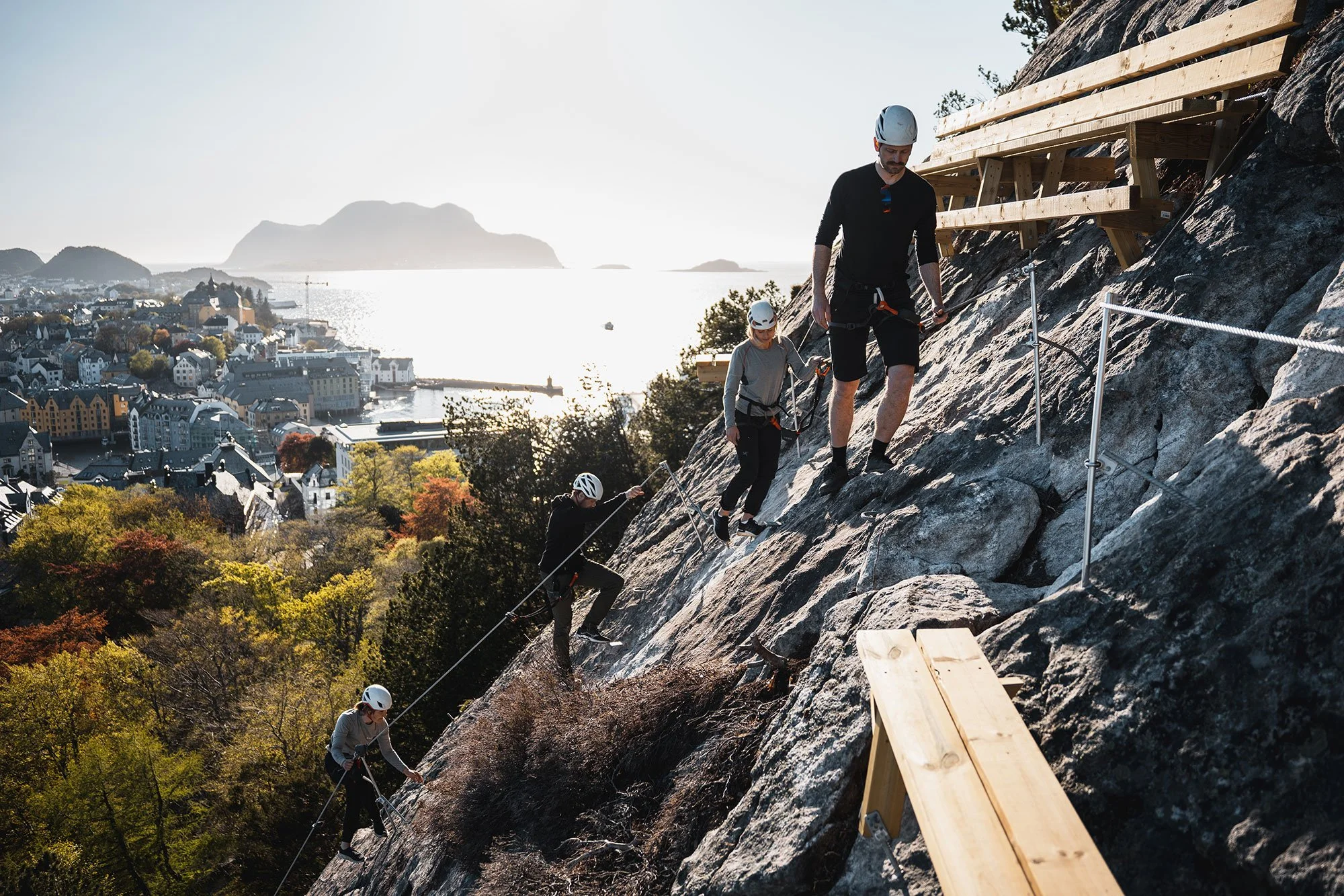 Explore Norway. Group of people climbing a steep rocky cliff with safety ropes and helmets, with a scenic coastal town and water in the background.