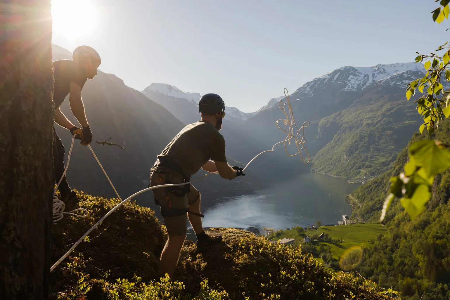 Explore Norway. Two men climbing a mountain, one of them is throwing a rope to the other, in a mountainous landscape with snow-capped peaks and a lake below, during sunset.