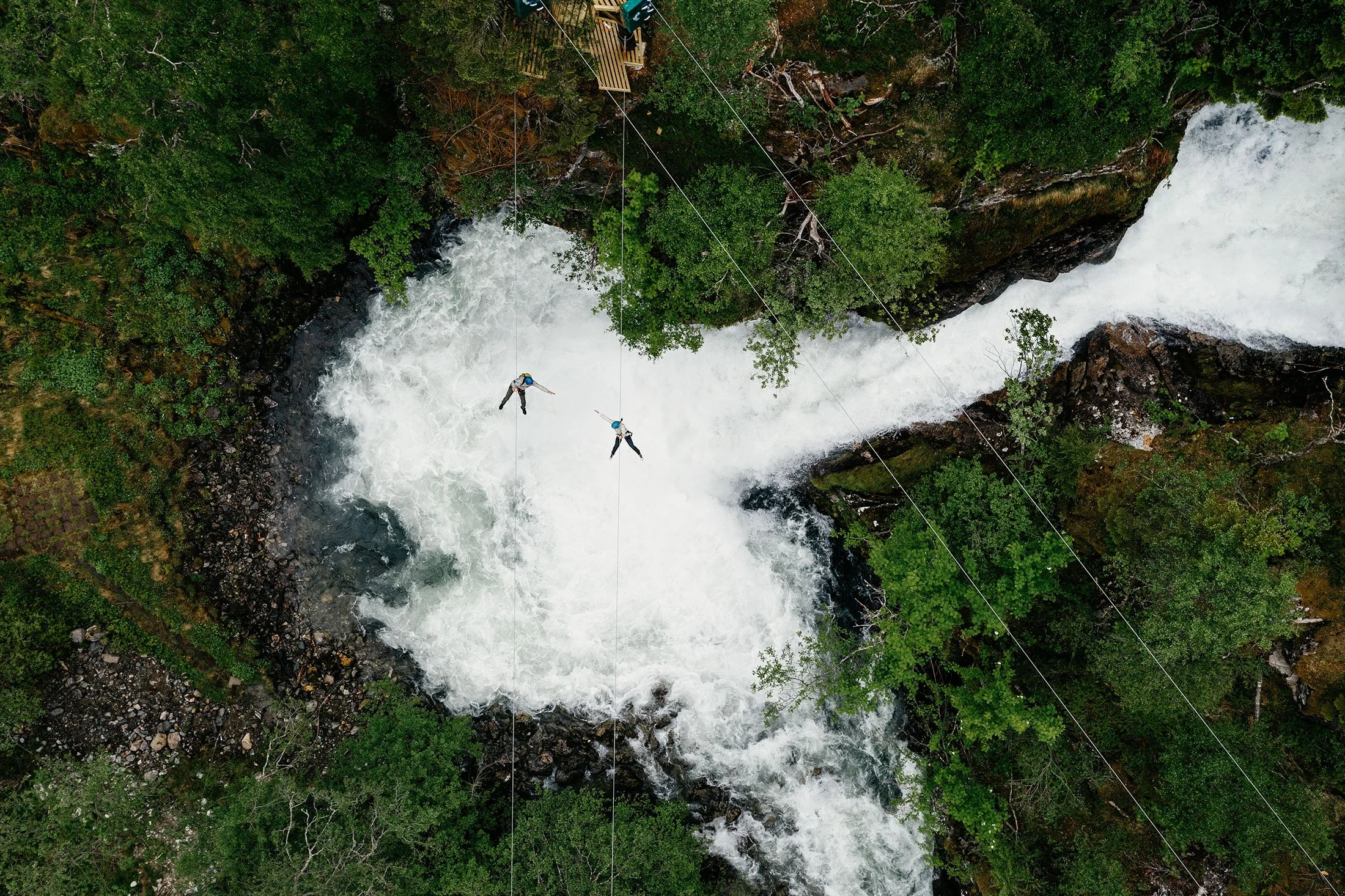 Two people zip-lining over a rushing river surrounded by lush green trees.