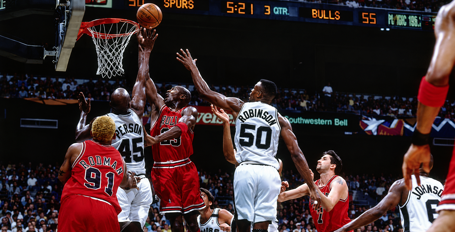 Basketball players in red and white uniforms compete near the basket during a game, with the crowd visible in the background.