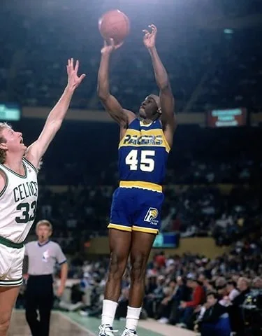 Basketball player wearing a blue and yellow uniform with the number 45 making a shot, while an opposing player reaching up to block in a crowded indoor court.