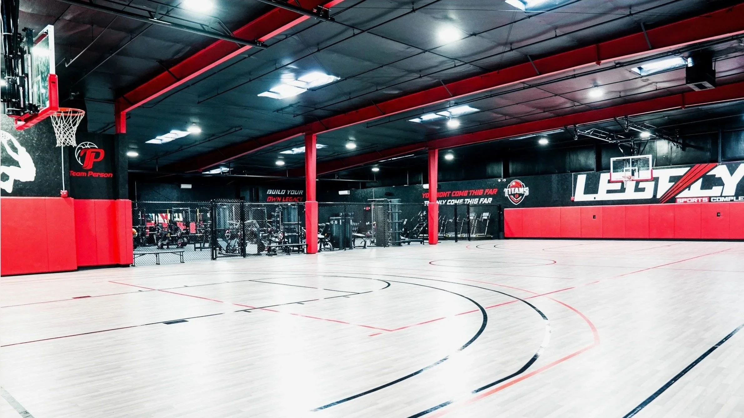 An empty indoor basketball court with red and black decor, including a basketball hoop, gym equipment area, and motivational slogans on the wall.