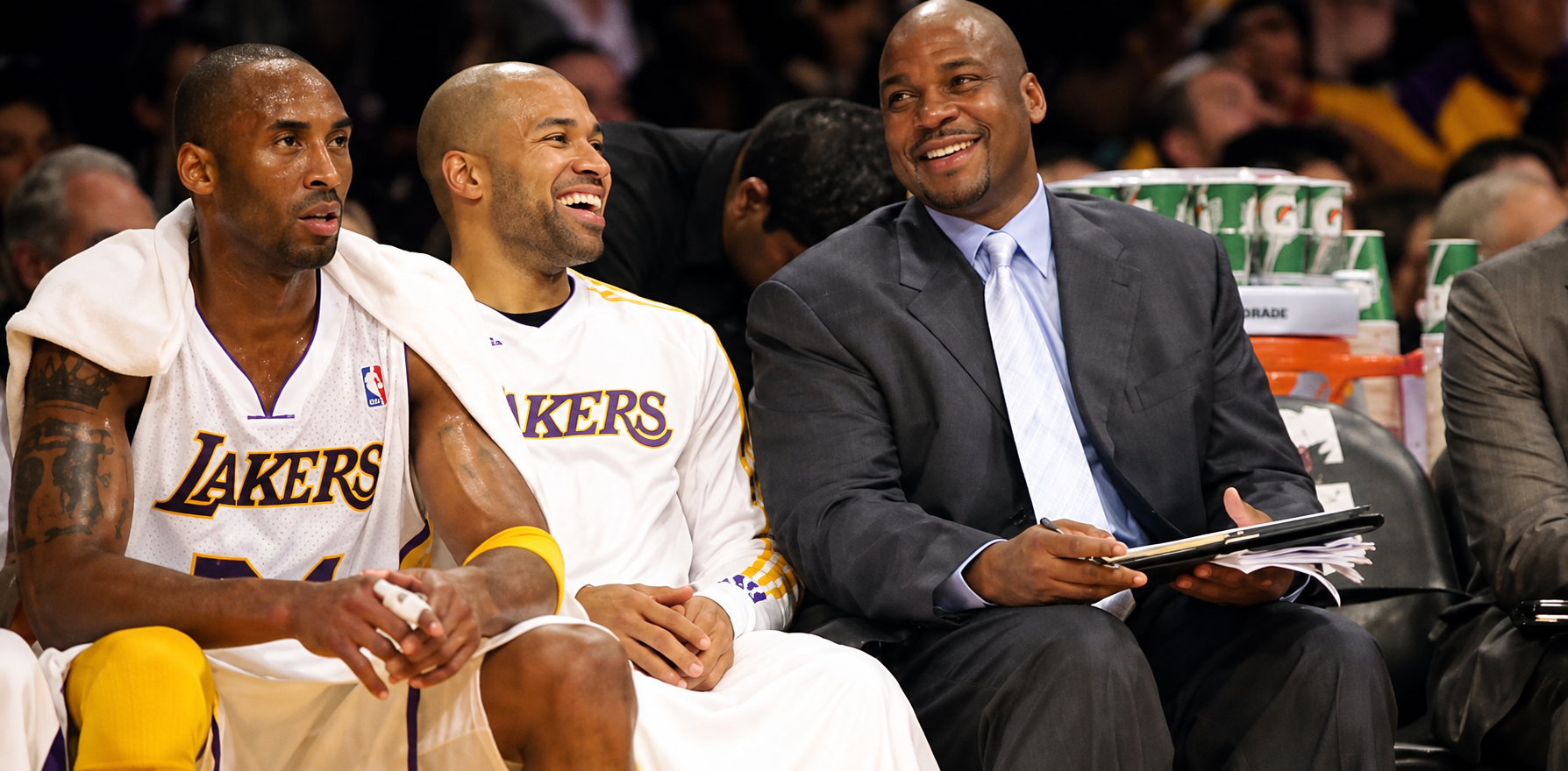 Two Los Angeles Lakers basketball players in white jerseys with purple and yellow accents sitting on the bench, smiling and talking. A man in a gray suit with a light blue shirt and tie, holding a clipboard, also sitting on the bench.