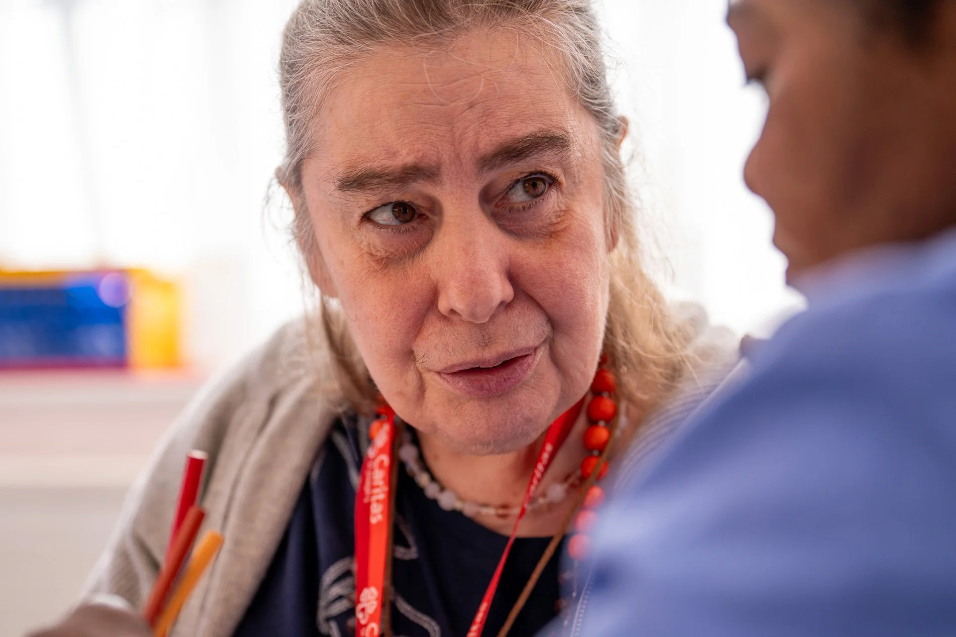 An elderly woman with gray hair, wearing a gray sweater, red necklace, and a red lanyard, engaged in a conversation with a person whose face is not visible.