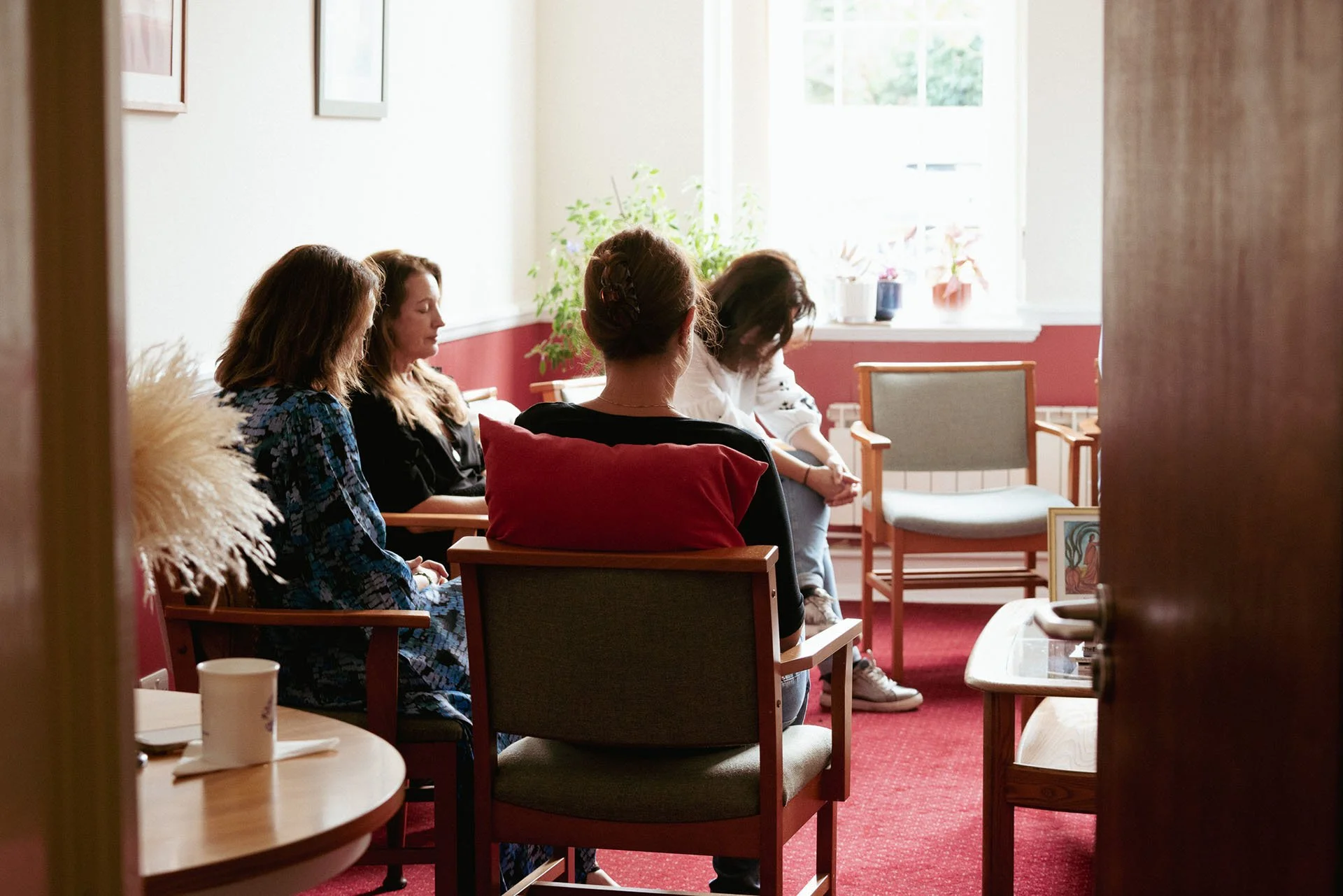Four women sitting in a circle with eyes closed, participating in a prayer session in a cozy room with natural light, red carpet, and potted plants.
