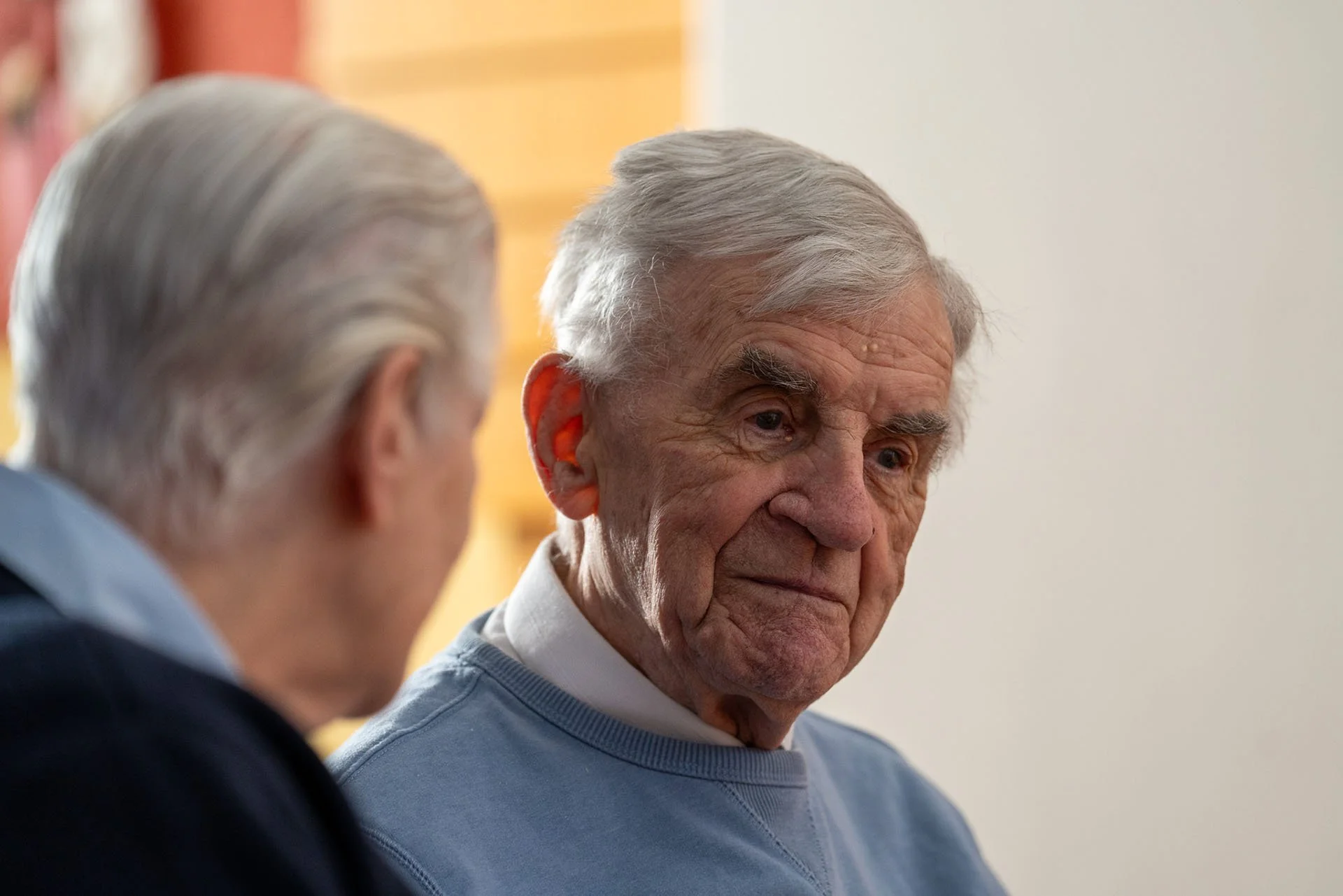Two elderly men having a conversation, one with gray hair and a somber expression, the other with blurred features, in a softly lit room.