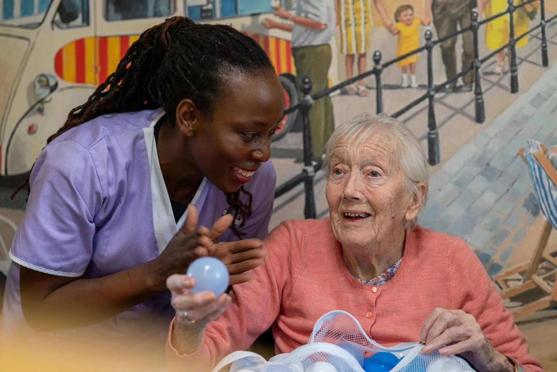 A caregiver and an elderly woman play with blue plastic balls, engaging in a joyful activity at a care facility with a colorful mural background.