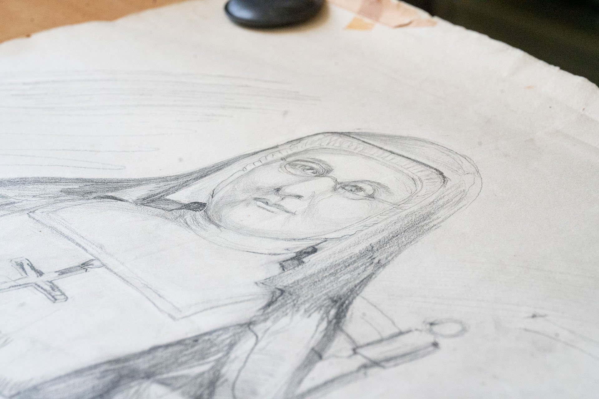 A detailed pencil sketch of a person's face on a piece of paper, with a portrait within a framed picture and a cross symbol nearby.