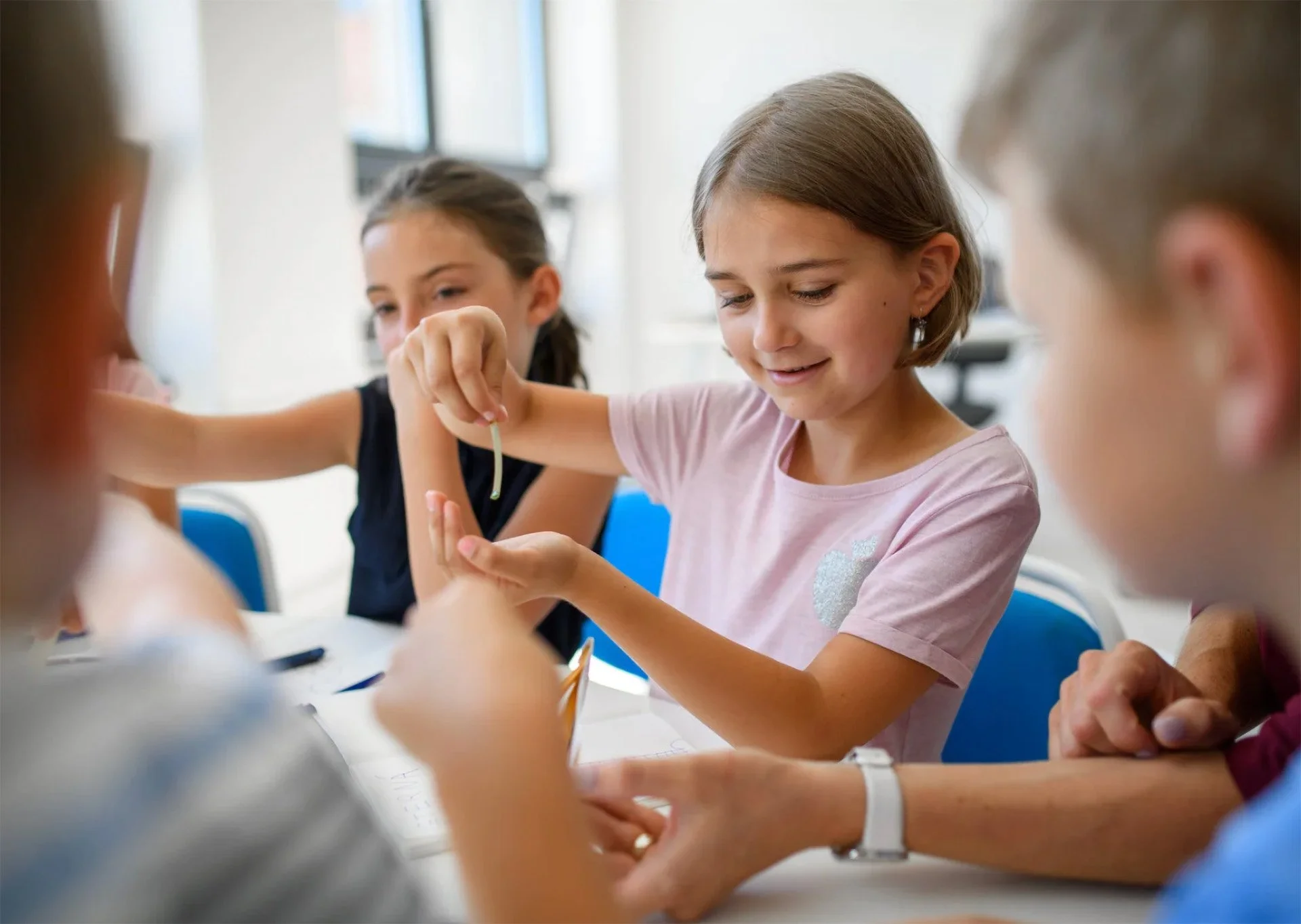 Children sitting at a table engaged in a science experiment, with one girl holding a small object and smiling.