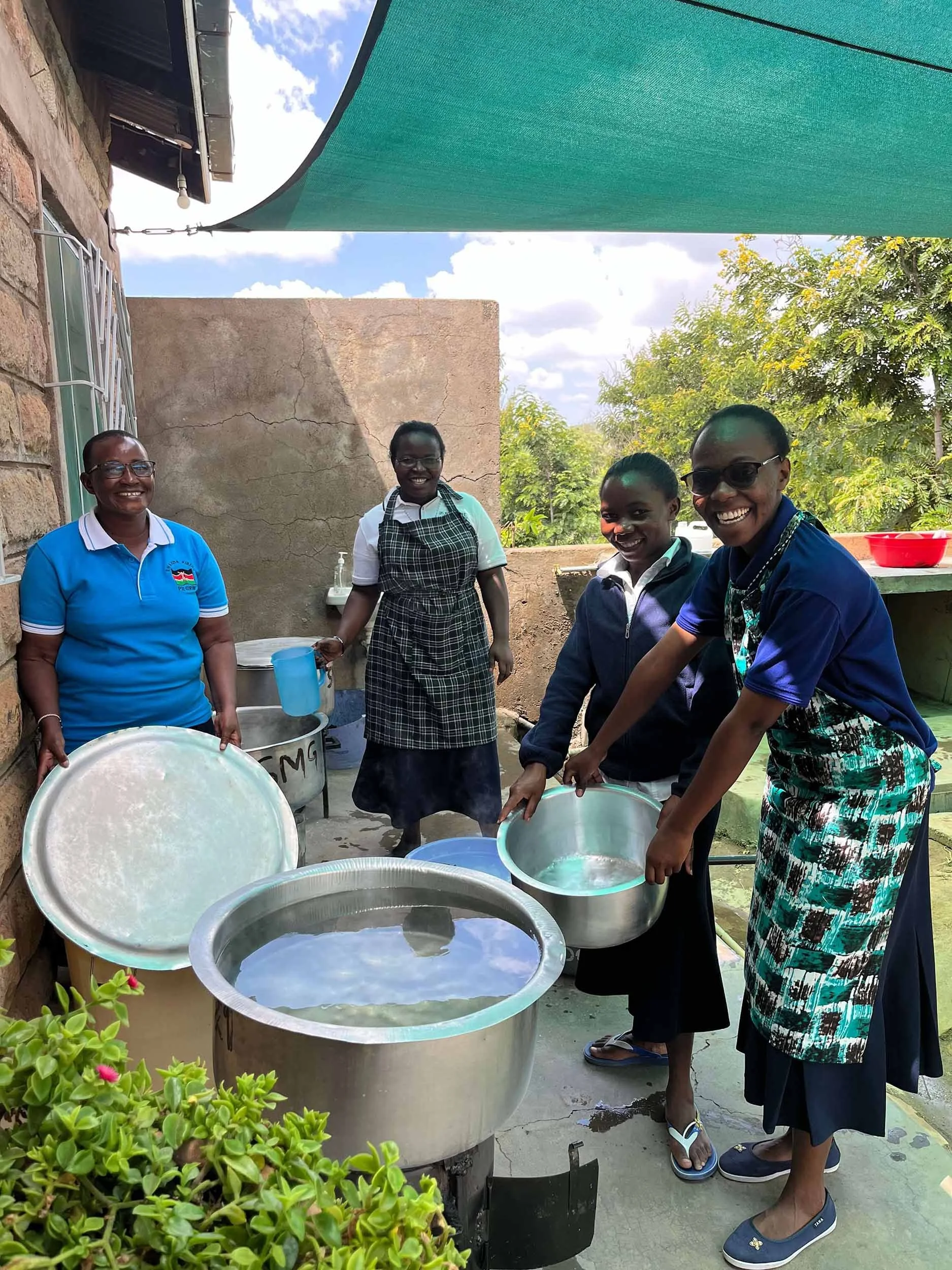 Four women are outdoors on a sunny day, washing large metal basins with soapy water. They are smiling and appear to be enjoying the activity. One woman is holding a blue measuring cup, and others are handling the basins. There is greenery and trees in the background, with a shaded canopy overhead.