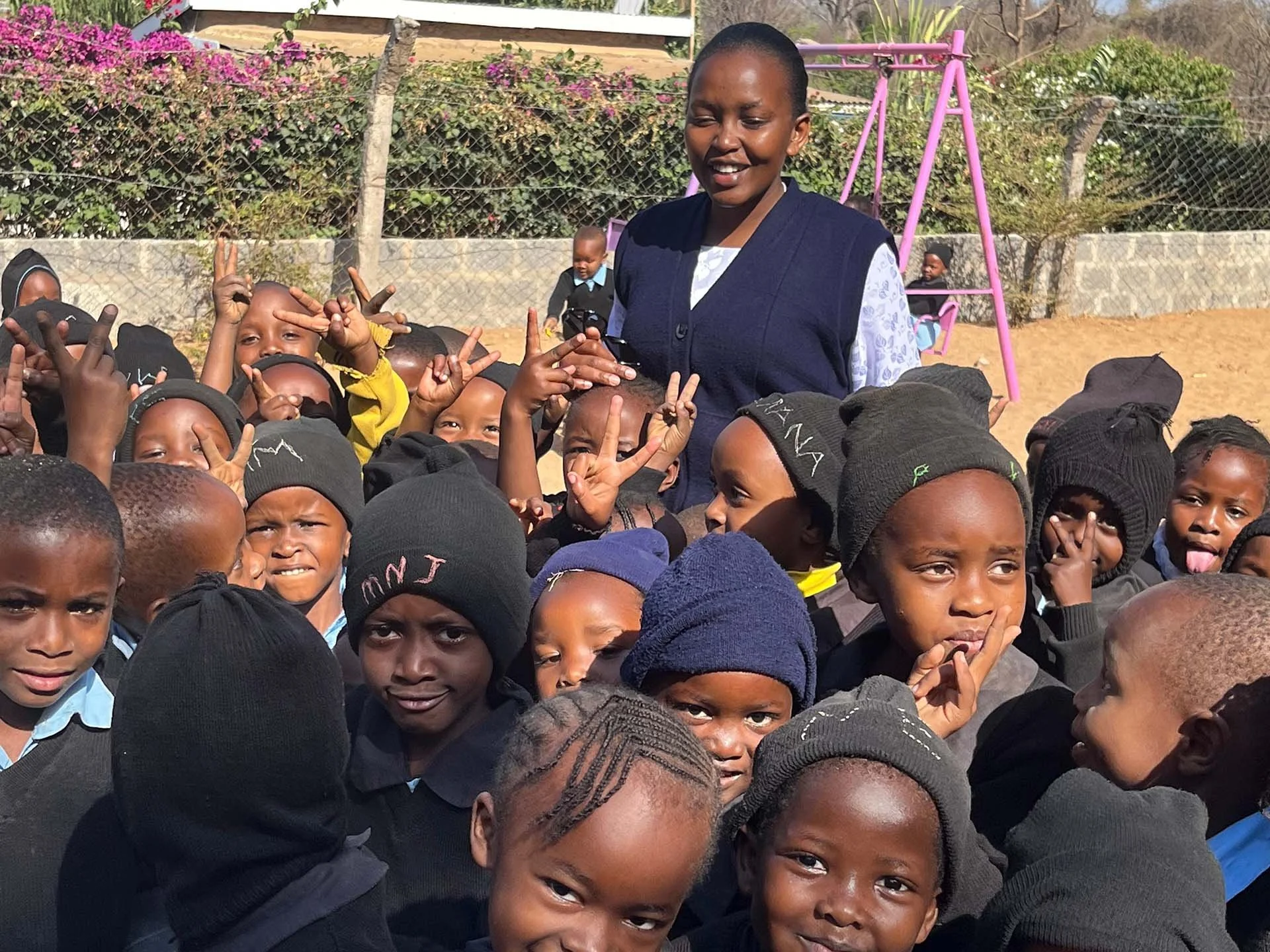 A group of children outdoors, a nun standing among them, some making peace signs and other gestures, with a pink swing set and a garden in the background.