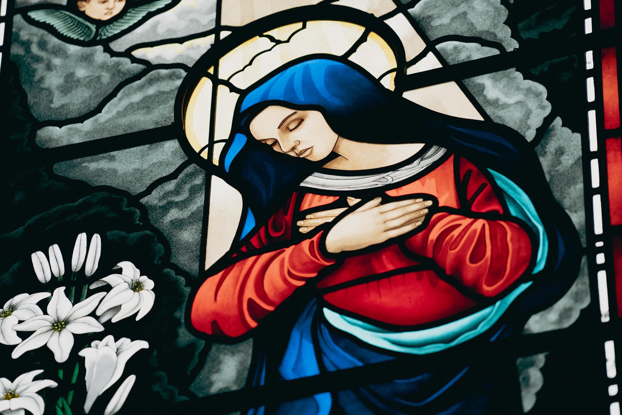 Stained glass depiction of the Virgin Mary with a halo, wearing a blue head covering and a red robe, with her hands crossed over her chest, set against a cloudy background with white lilies in the foreground.