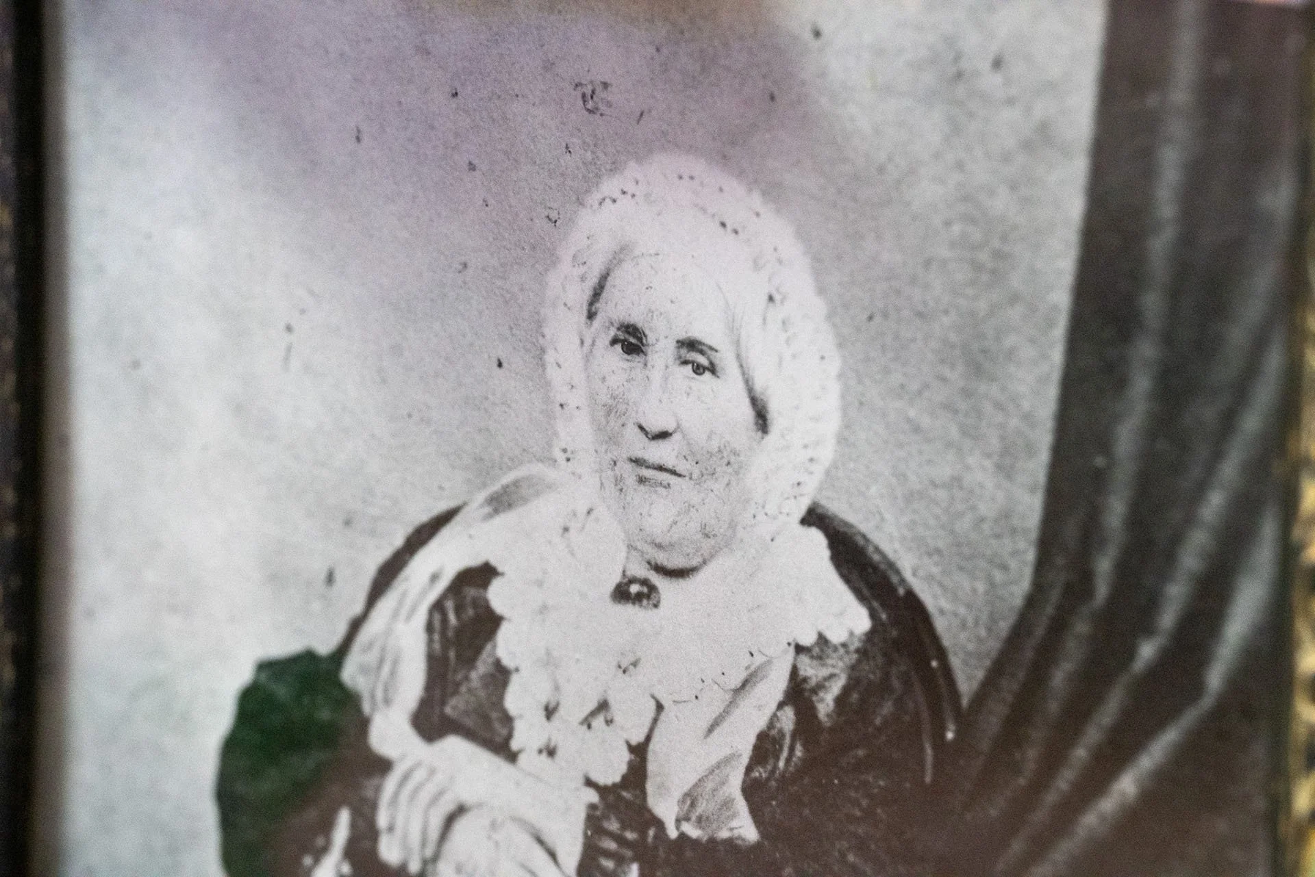 A black-and-white portrait of an elderly woman with a serious expression. She is dressed in period clothing with a lace collar and has her hair styled back. The background is plain and textured.