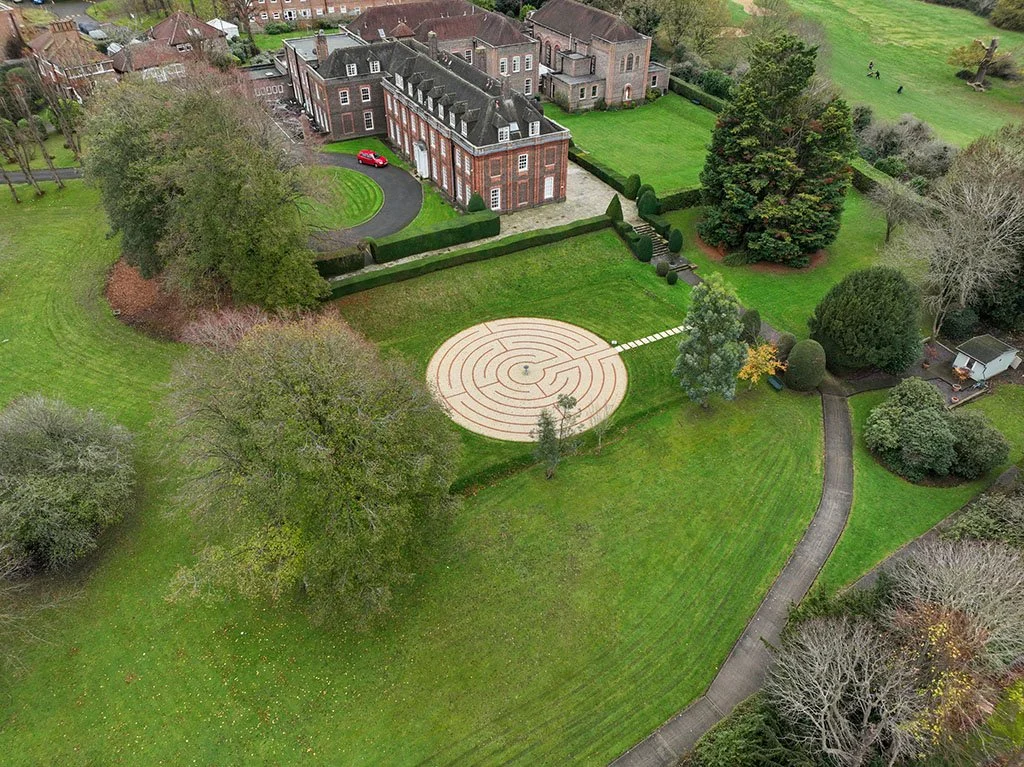 Aerial view of a large estate with a circular labyrinth garden, green lawns, trees, and a multi-story brick house with a curved driveway with a red car parked.