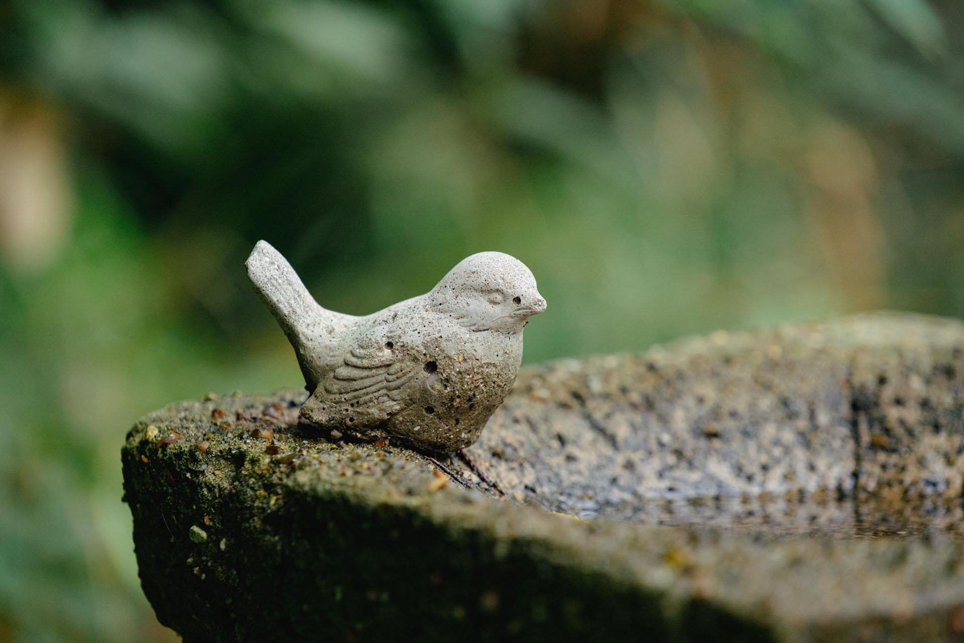 A small bird-shaped stone sculpture resting on a moss-covered rock with a blurred green background.