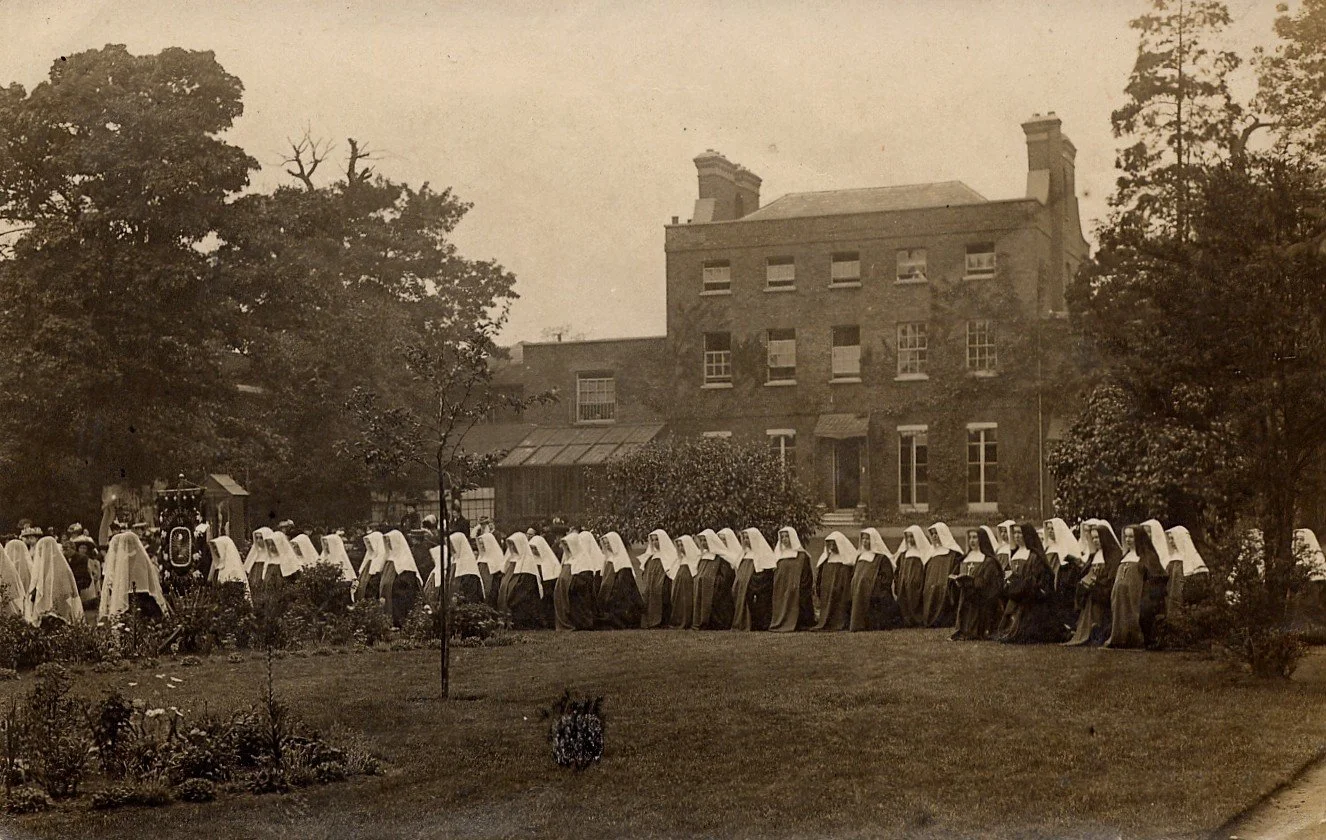 Vintage black and white photo of Catholic nuns in habit and veil walking in a line outside a large brick building with trees and garden in the background.