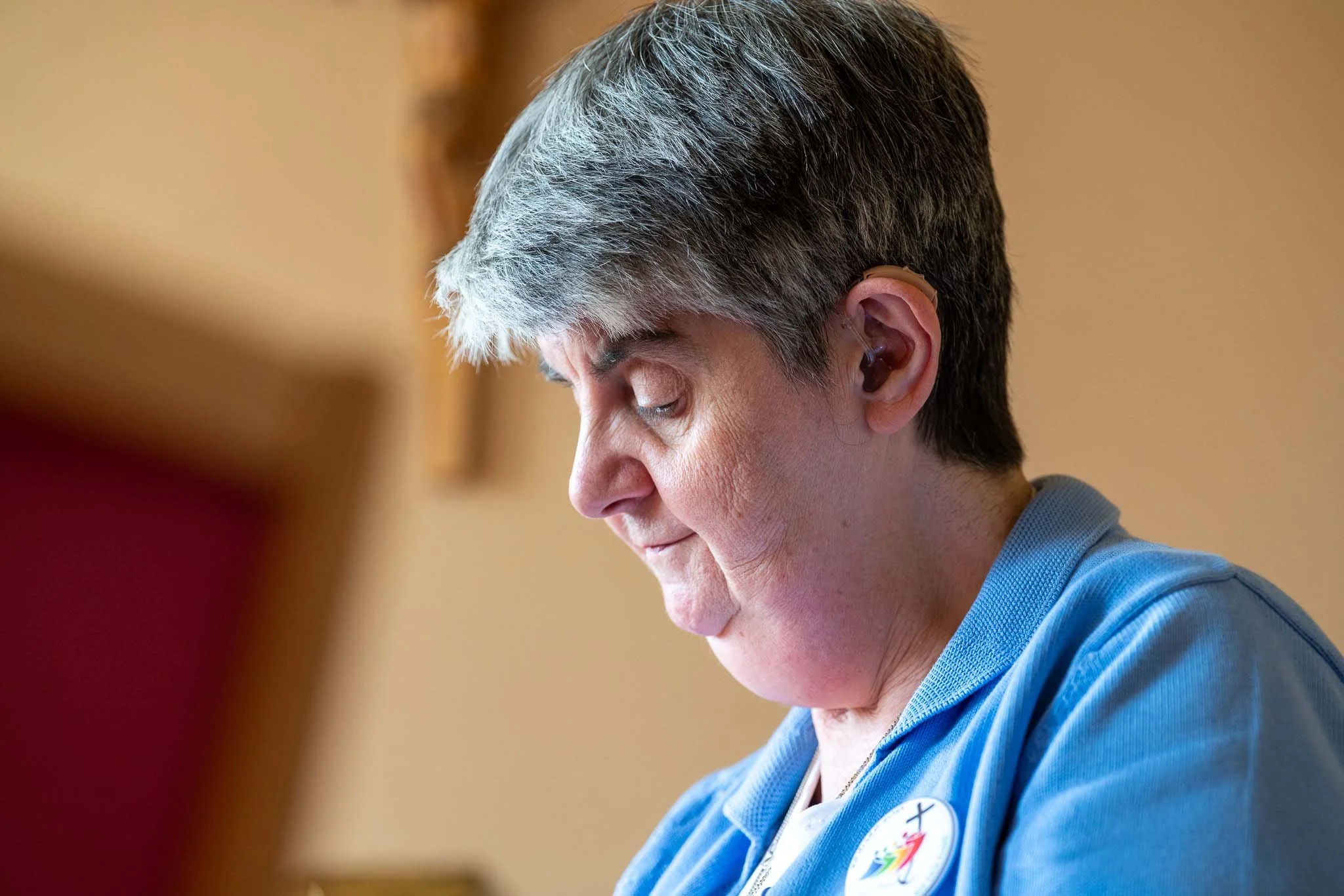 A woman with short gray hair wearing a blue top, with her eyes closed and head tilted downward, appears to be in a moment of reflection or prayer.