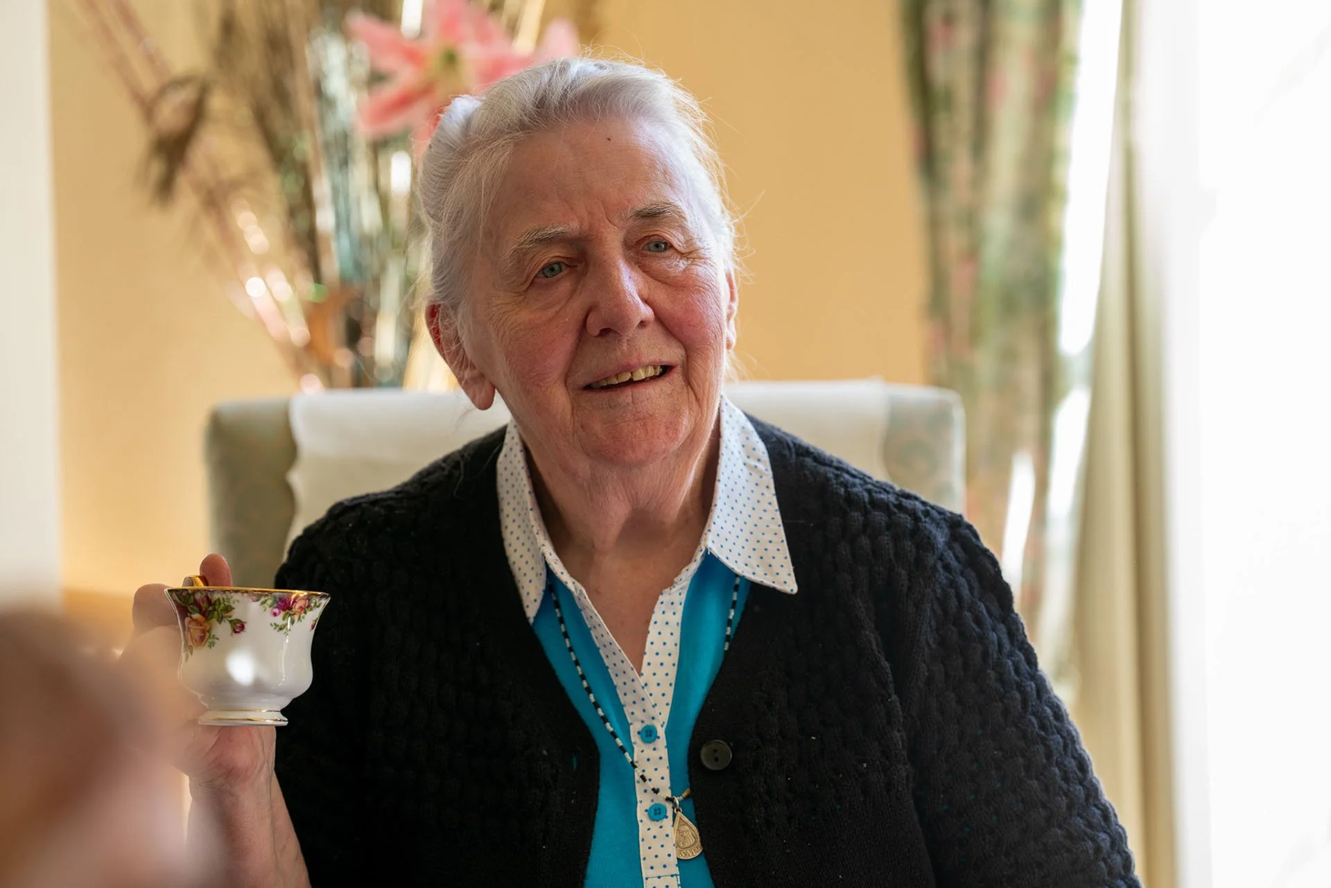 An elderly woman with white hair, wearing a black knitted cardigan, holding a teacup with a floral pattern, smiling in a well-lit room with floral curtains.